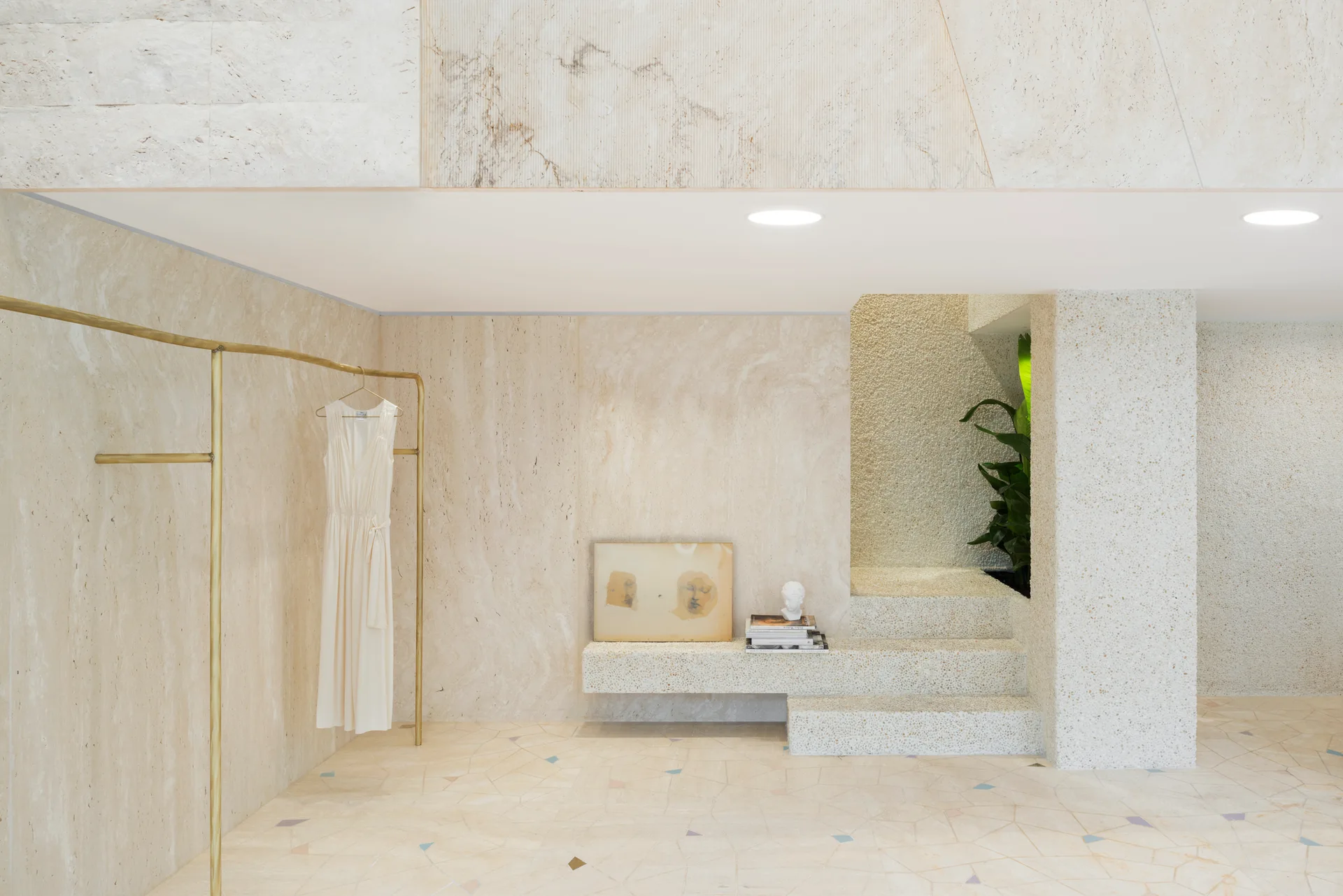 A minimalist stone interior featuring cream travertine walls, a brass clothing rack, and a terrazzo staircase with an integrated display bench.