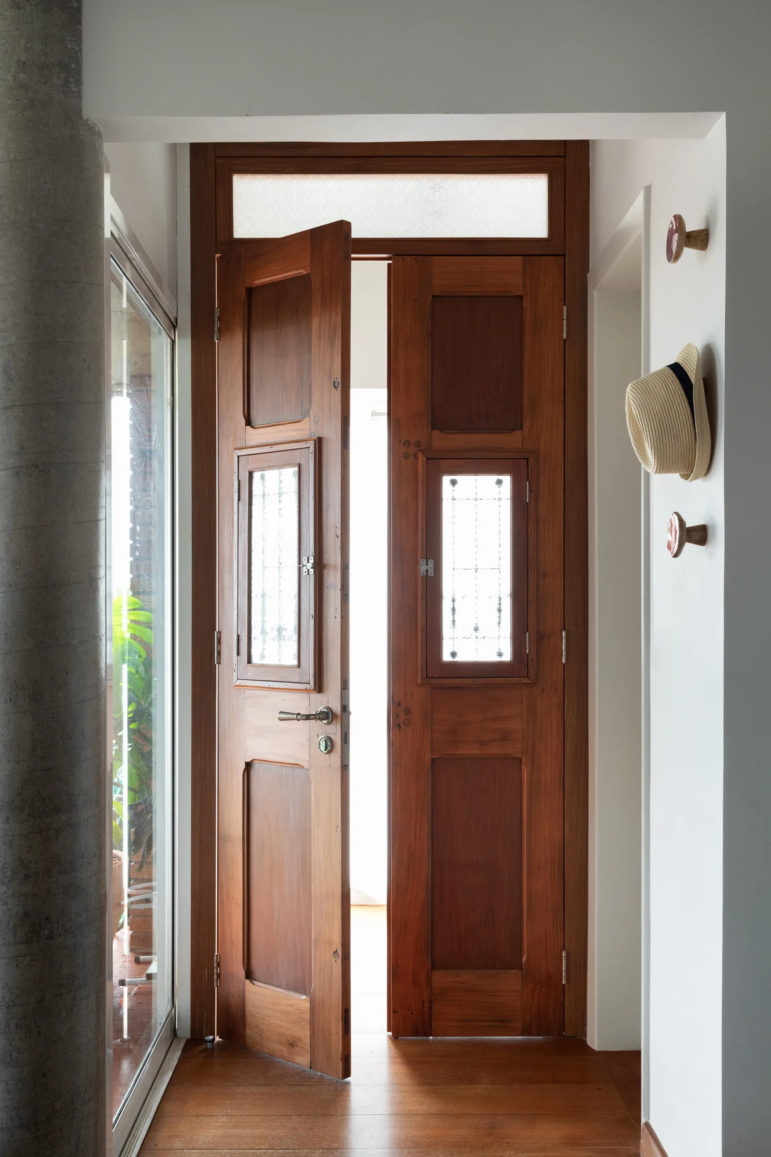 A warm wooden double door stands slightly open in a bright entryway with white walls, a concrete column, and wood flooring.