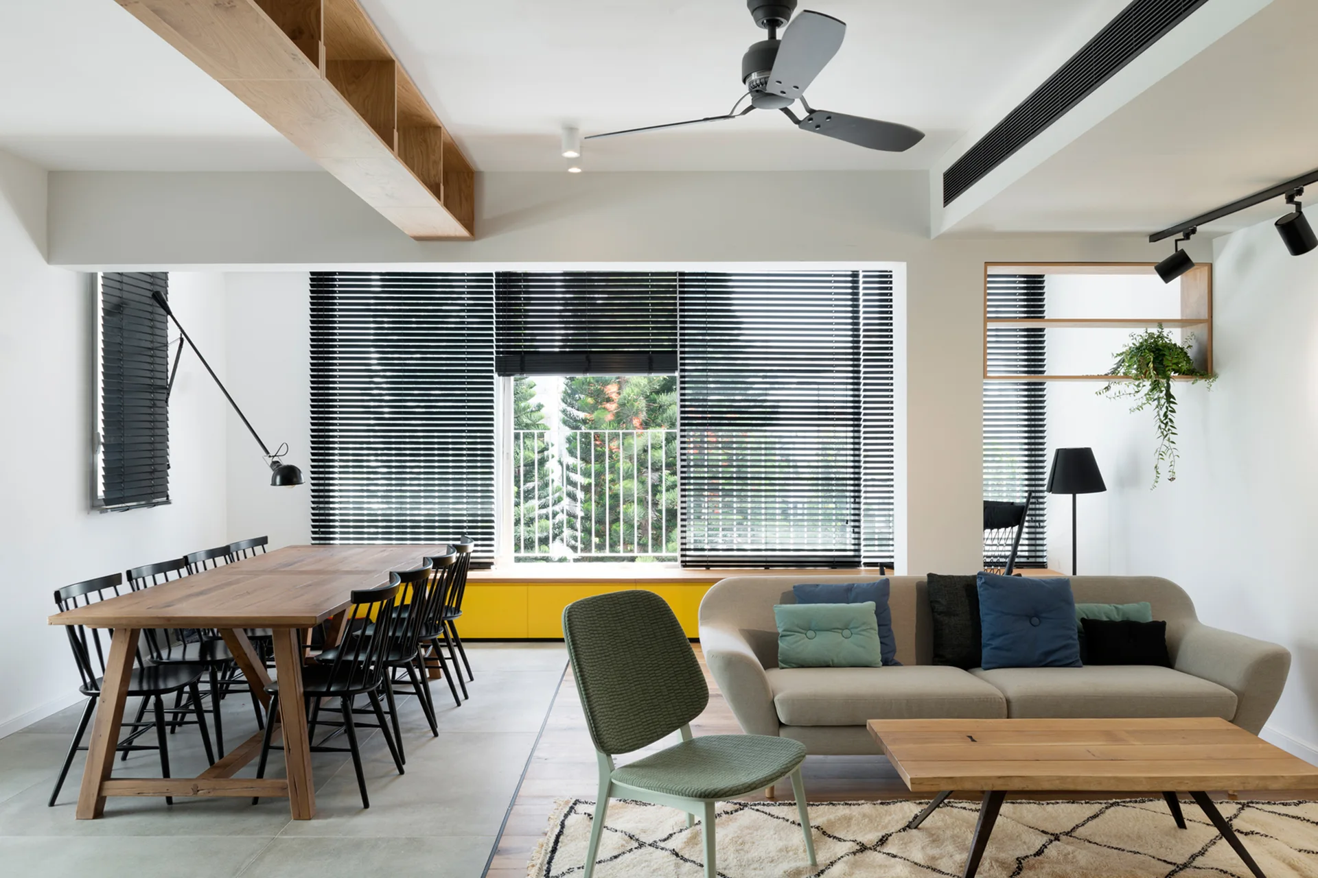 A modern open-concept space features a wooden dining table with black chairs alongside a grey sofa and a low yellow cabinet beneath black blinds.