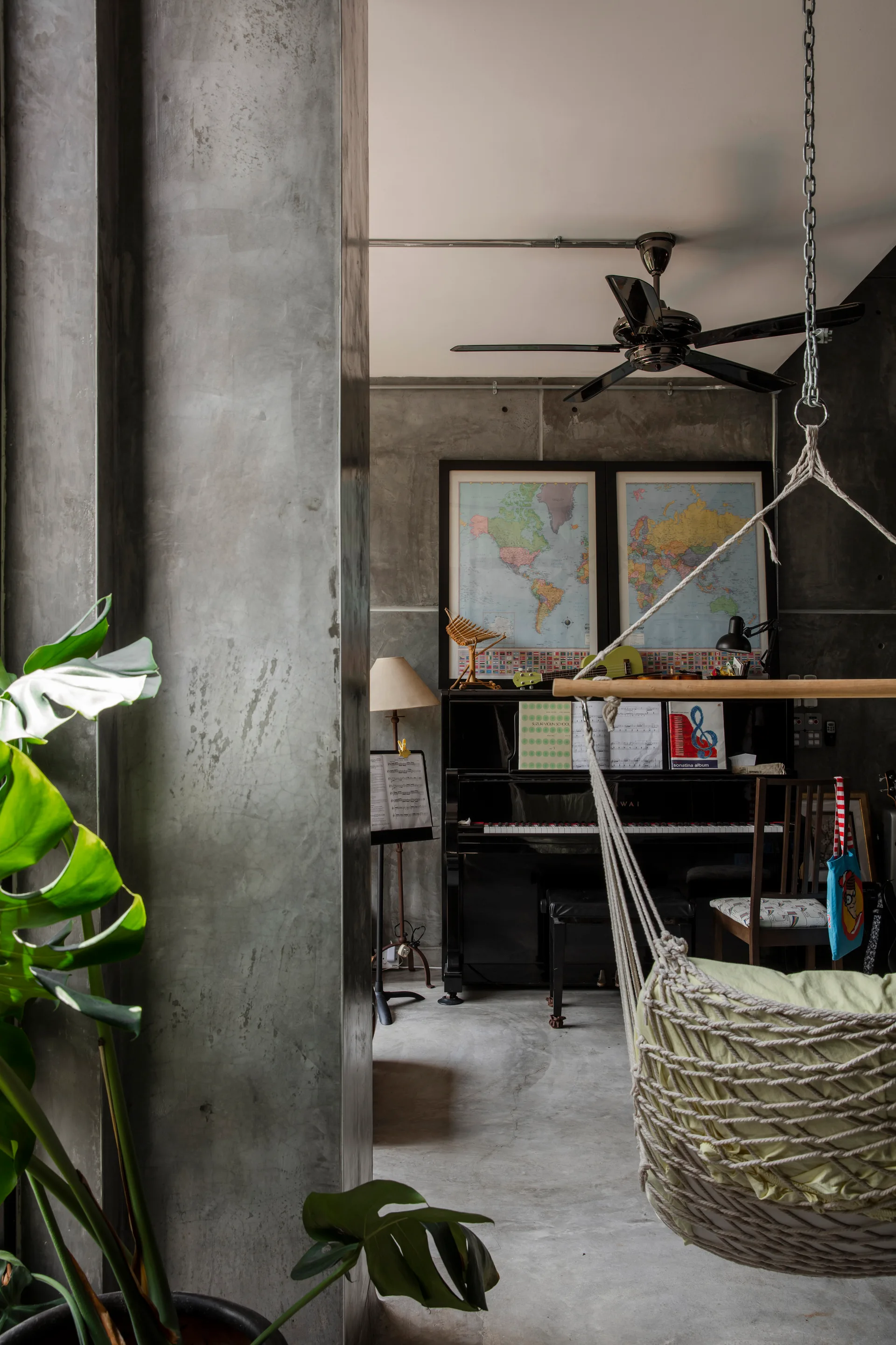 An upright piano sits against a gray concrete wall adorned with framed maps, beside a hanging woven swing chair and a large potted monstera plant.