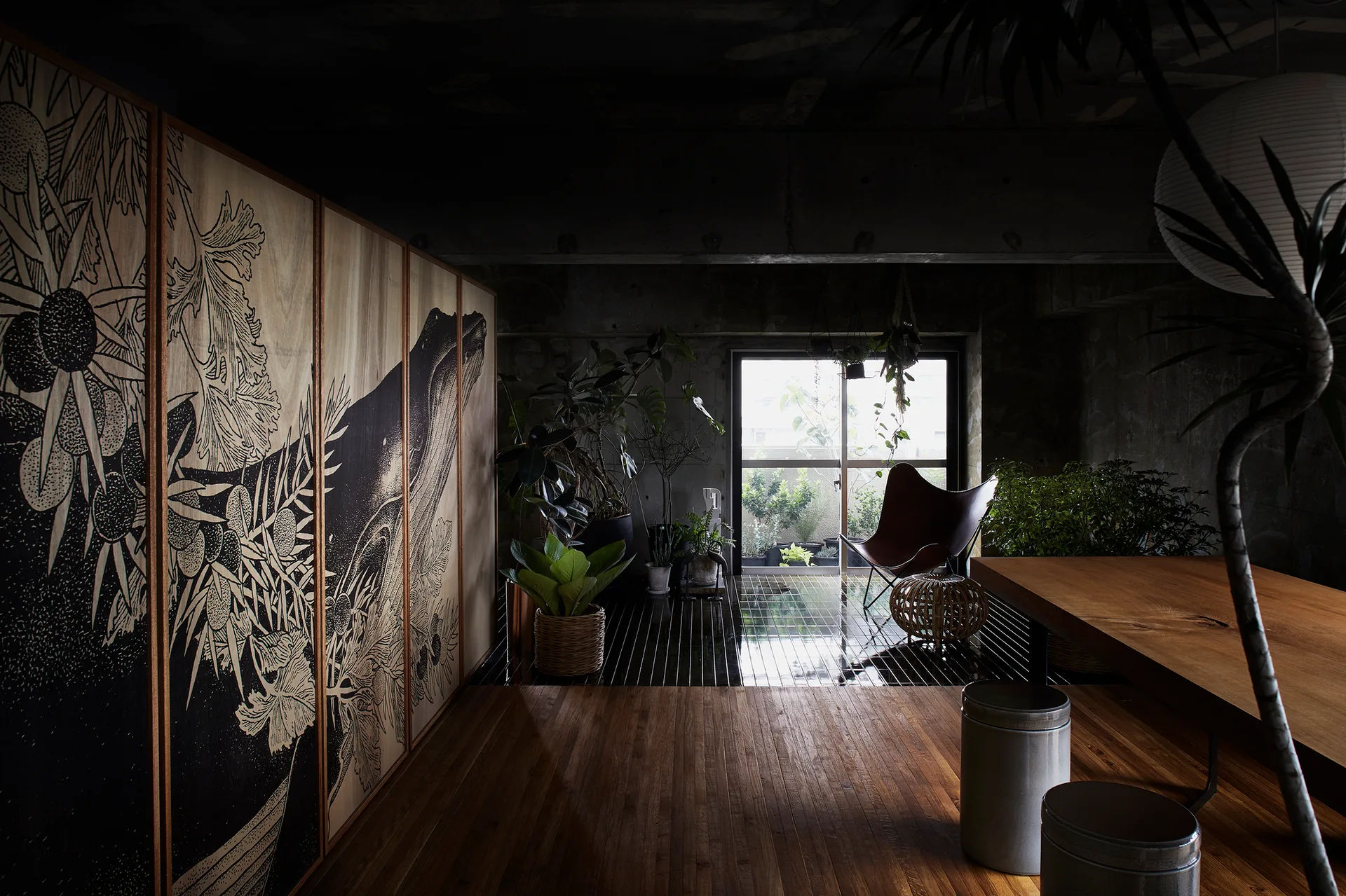 A moody living room features a large wood cabinet with botanical illustrations, concrete walls, potted plants, and a brown leather butterfly chair.