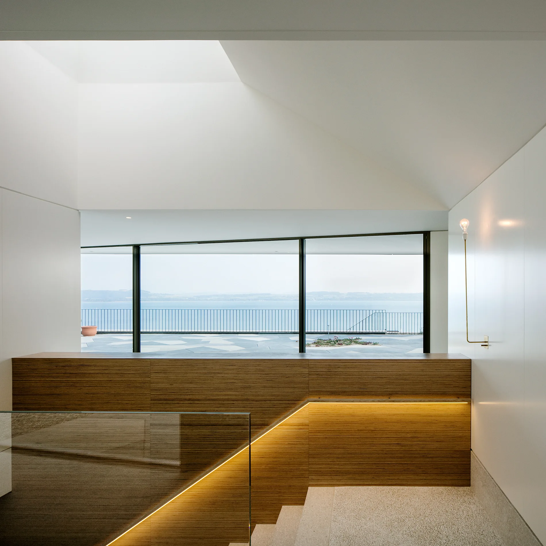 A minimalist staircase with light stone steps, wood paneling, an integrated warm LED handrail light, and a panoramic window overlooking water.