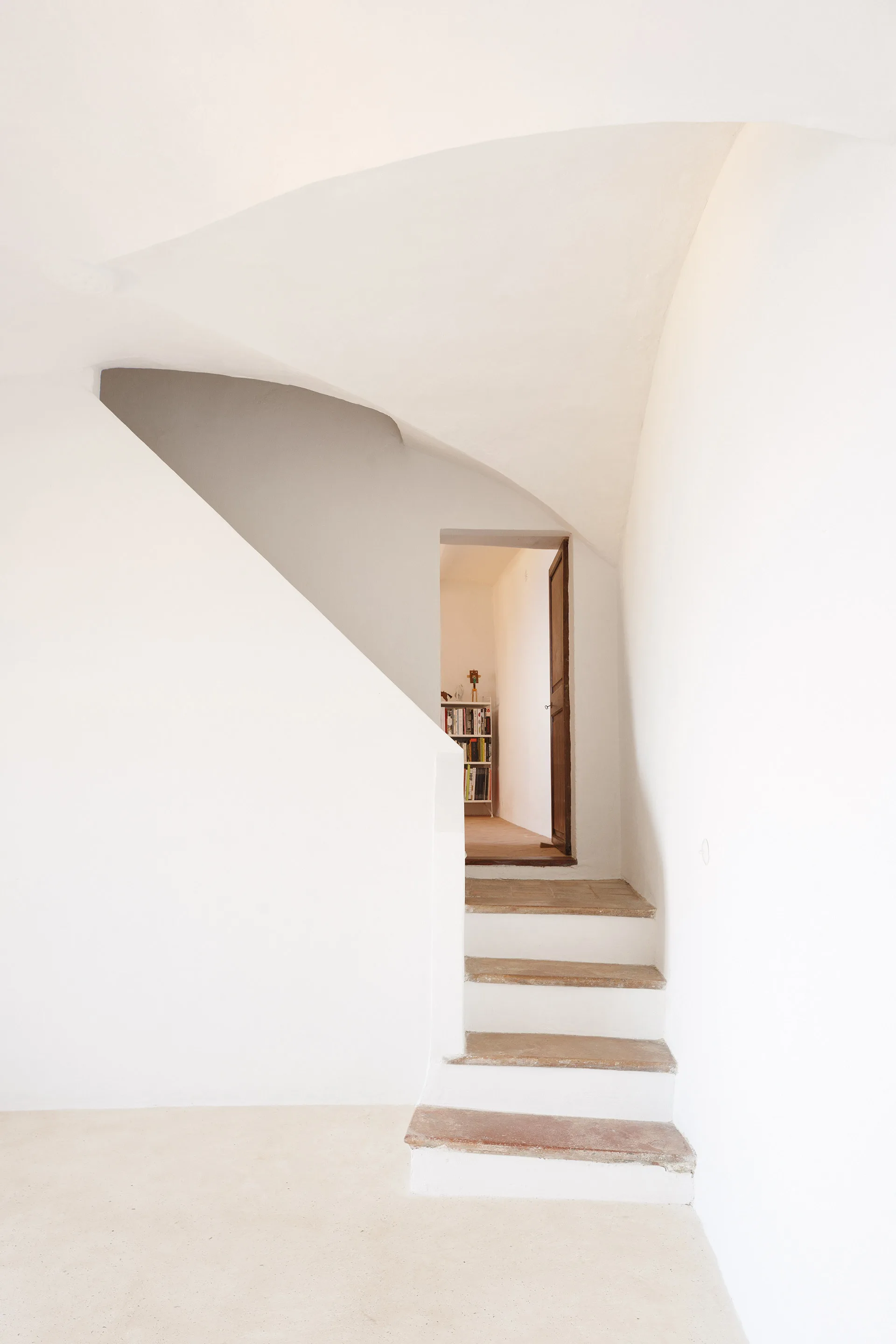 A minimalist white plaster staircase with natural stone treads leads up to an open wooden door under a vaulted ceiling.