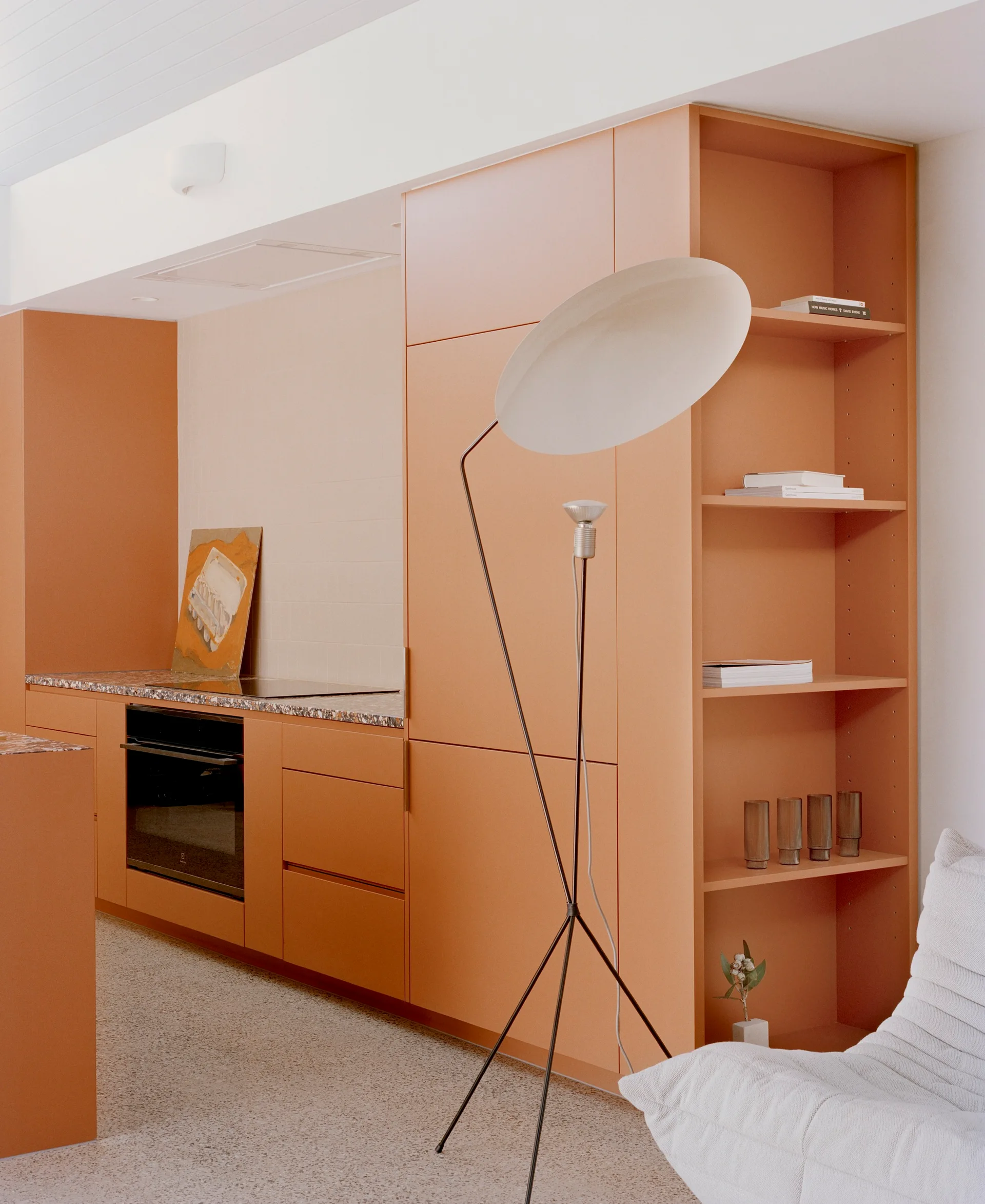 A modern kitchen featuring matte terracotta cabinetry with integrated open shelving, a speckled terrazzo countertop, and a white tripod floor lamp.