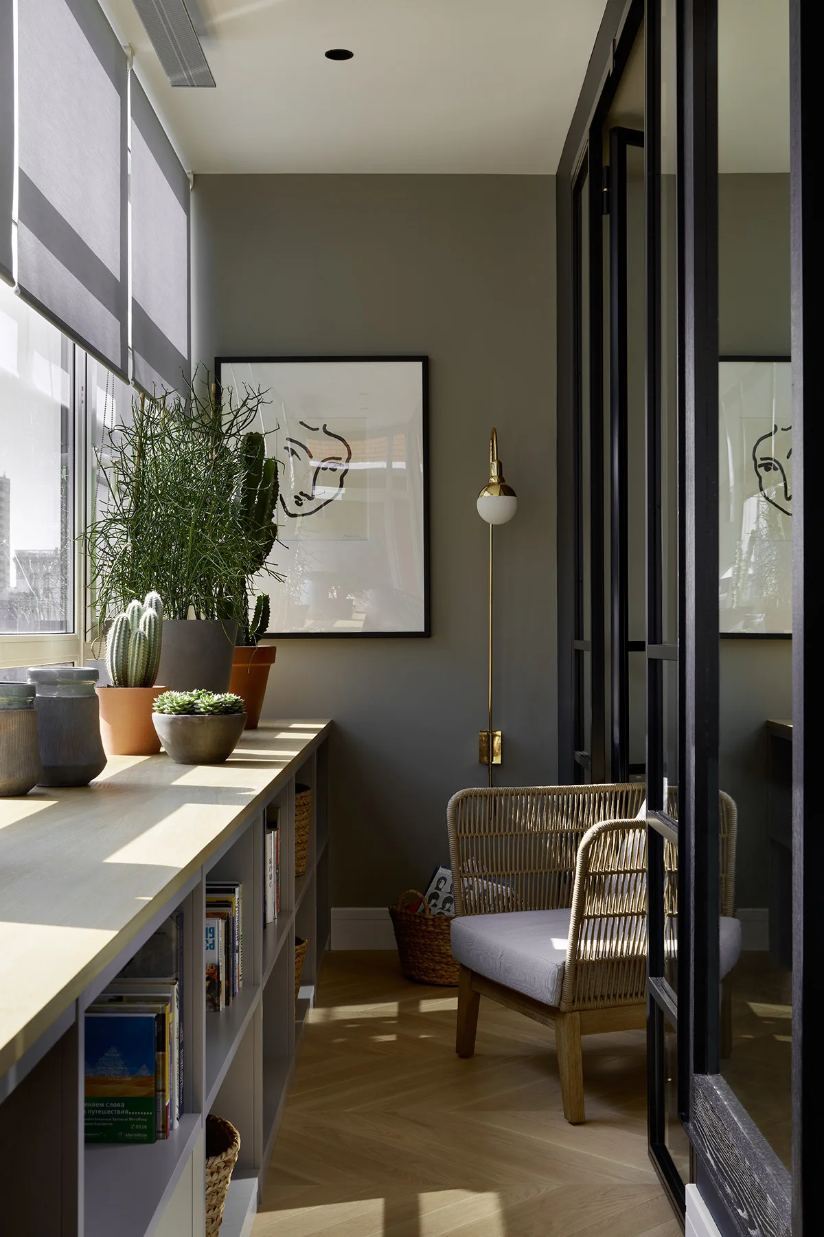 A sunlit reading nook features a wicker armchair, sage green walls, a low built-in bookcase under a window, and a black-framed glass partition.