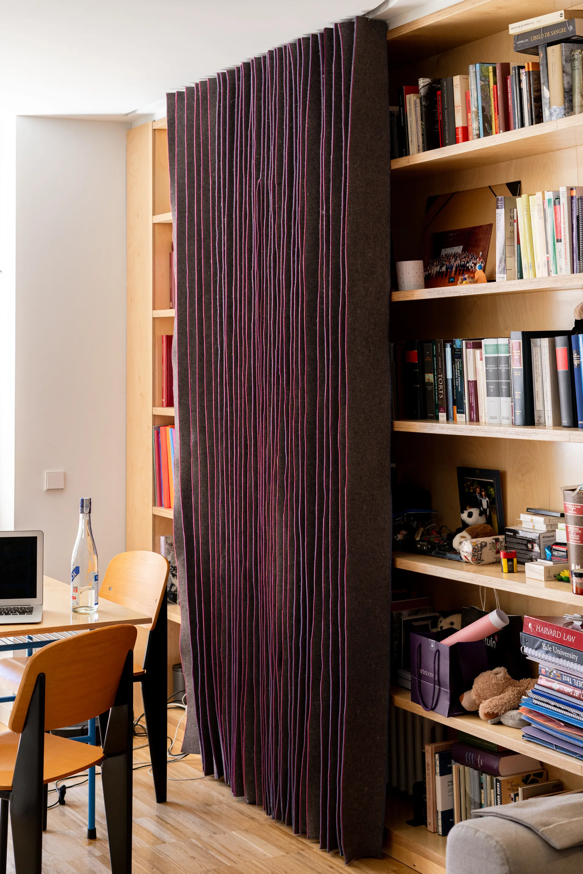 A light wood built-in bookshelf in a home office features a dark grey pleated fabric sliding screen alongside a wooden desk and chairs.