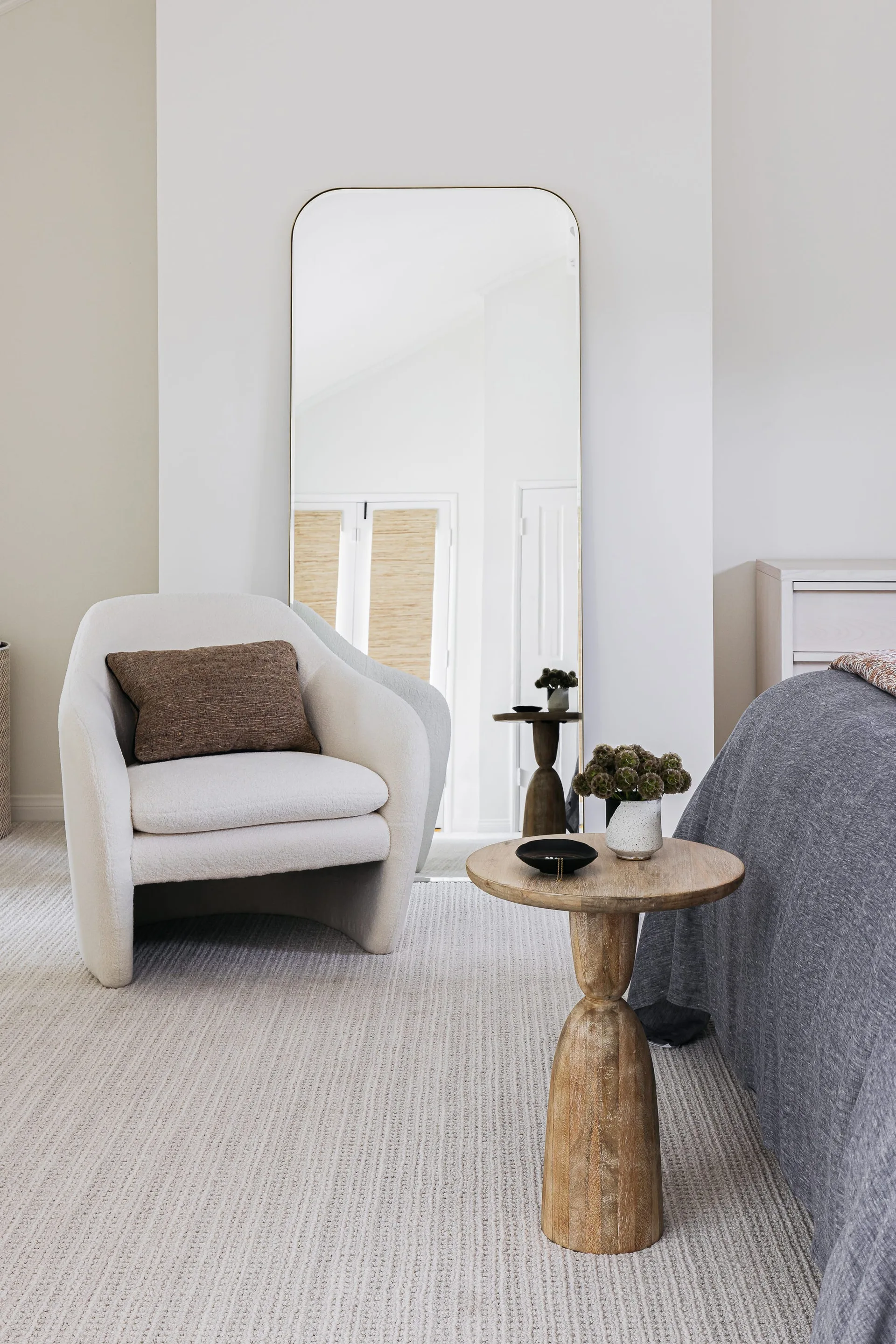 A bedroom corner features a curved white armchair, a tall arched floor mirror, and a round wooden pedestal side table on textured carpet.