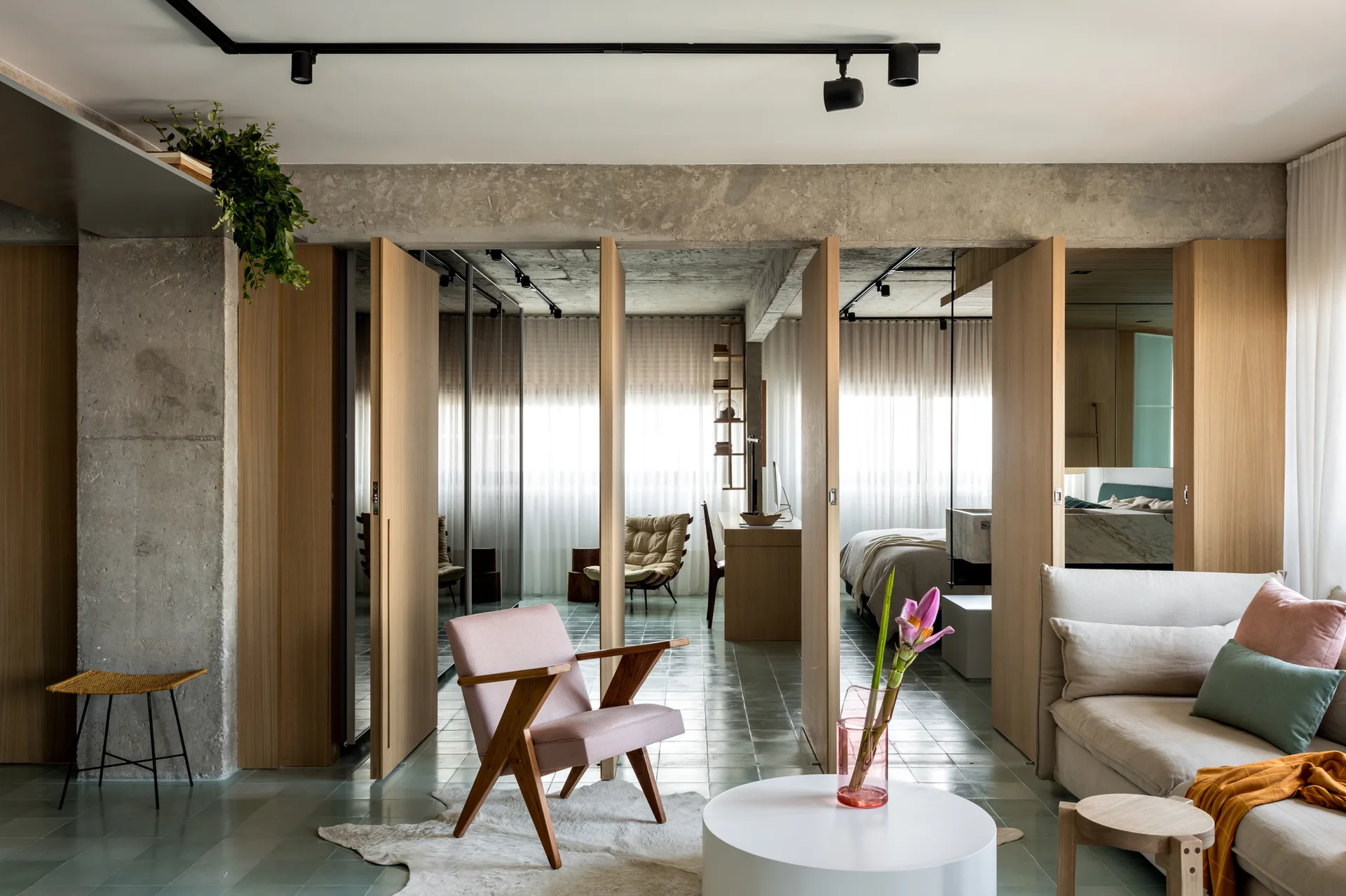 A modern living room features pivoting vertical wood partitions, an exposed concrete beam, a pink armchair, and a beige sofa on a tiled floor.