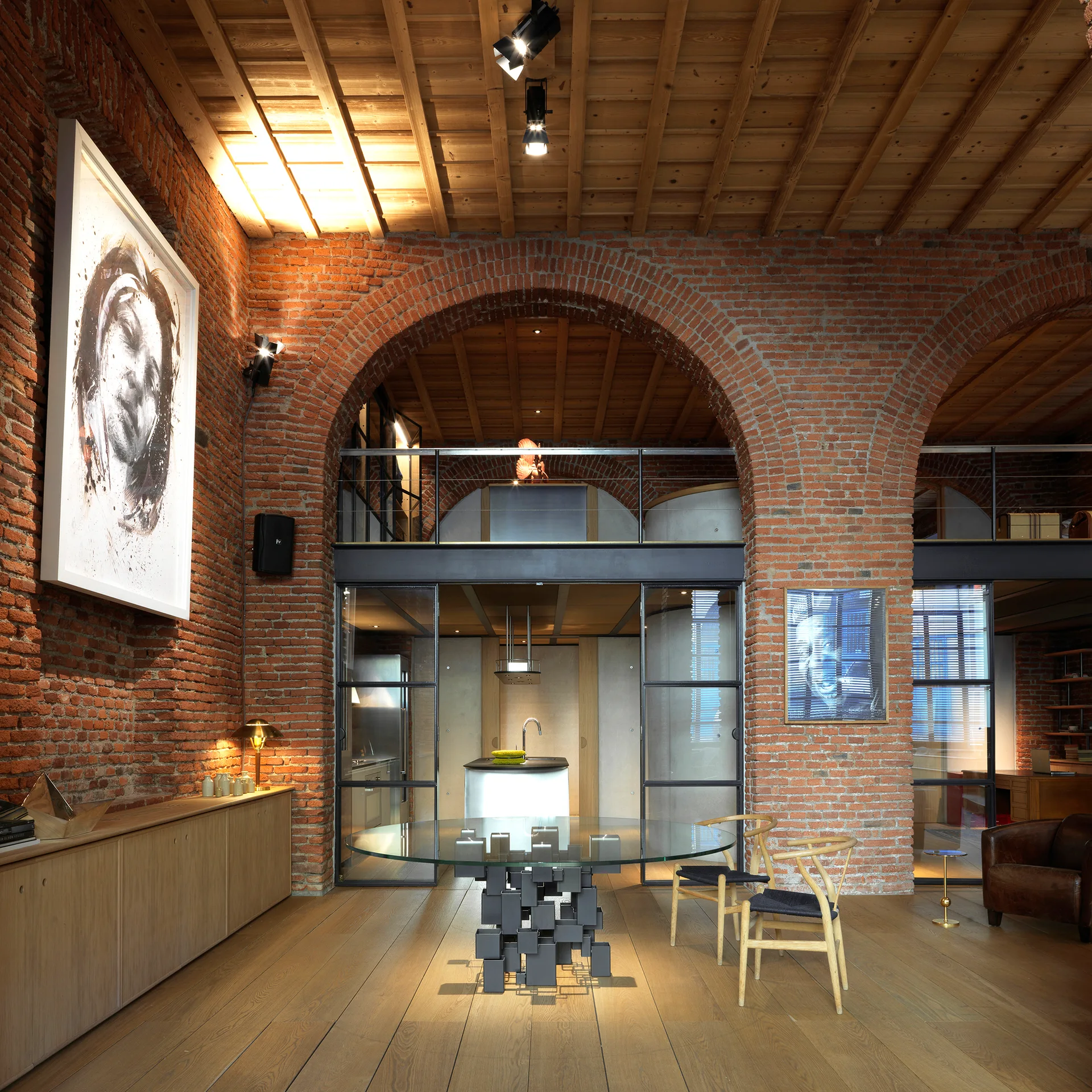 A loft dining space features exposed red brick arches, a round glass-topped table with wishbone chairs, and black-framed glass partition walls.