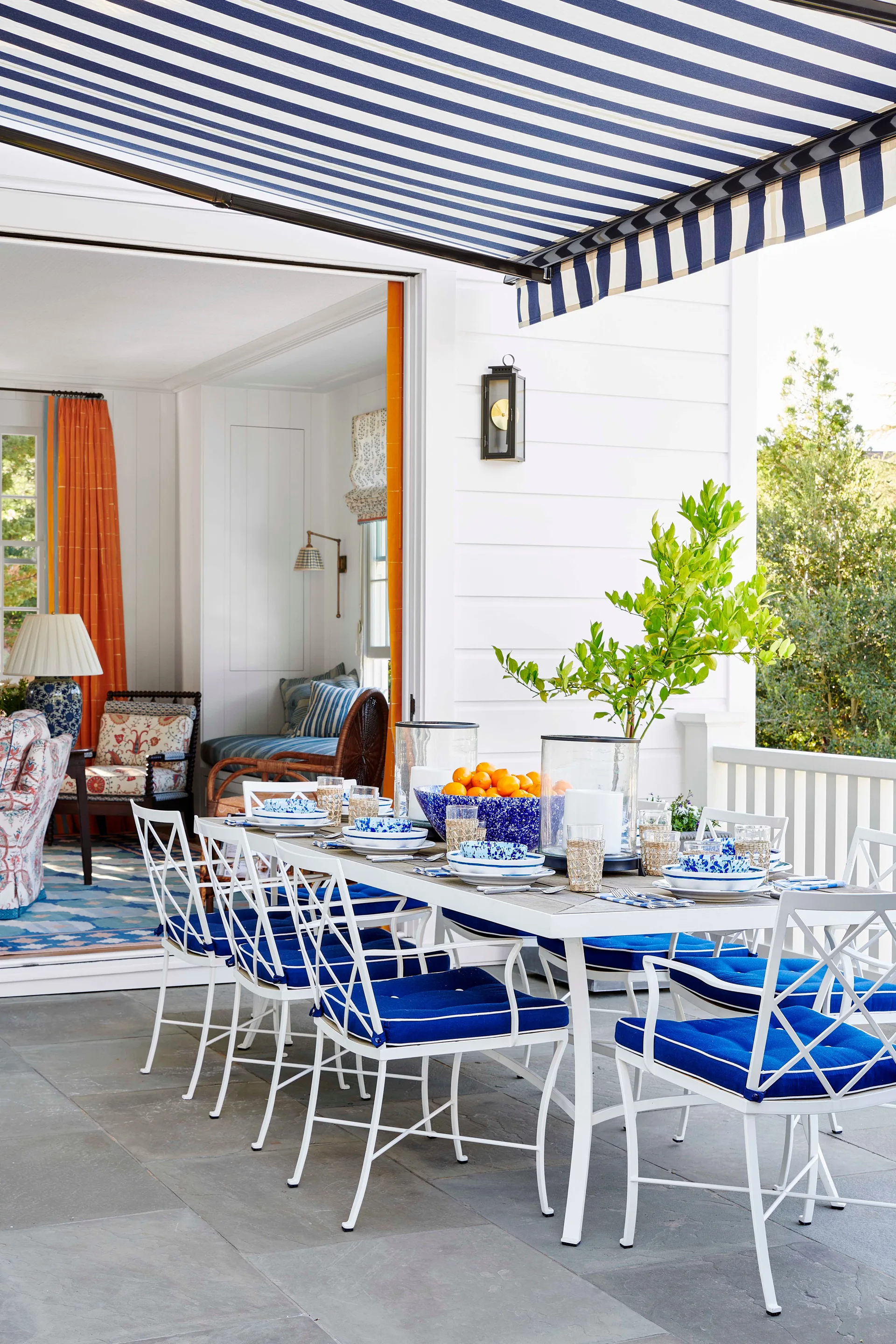 An outdoor dining table set with blue and white tableware sits under a striped awning next to an open doorway.