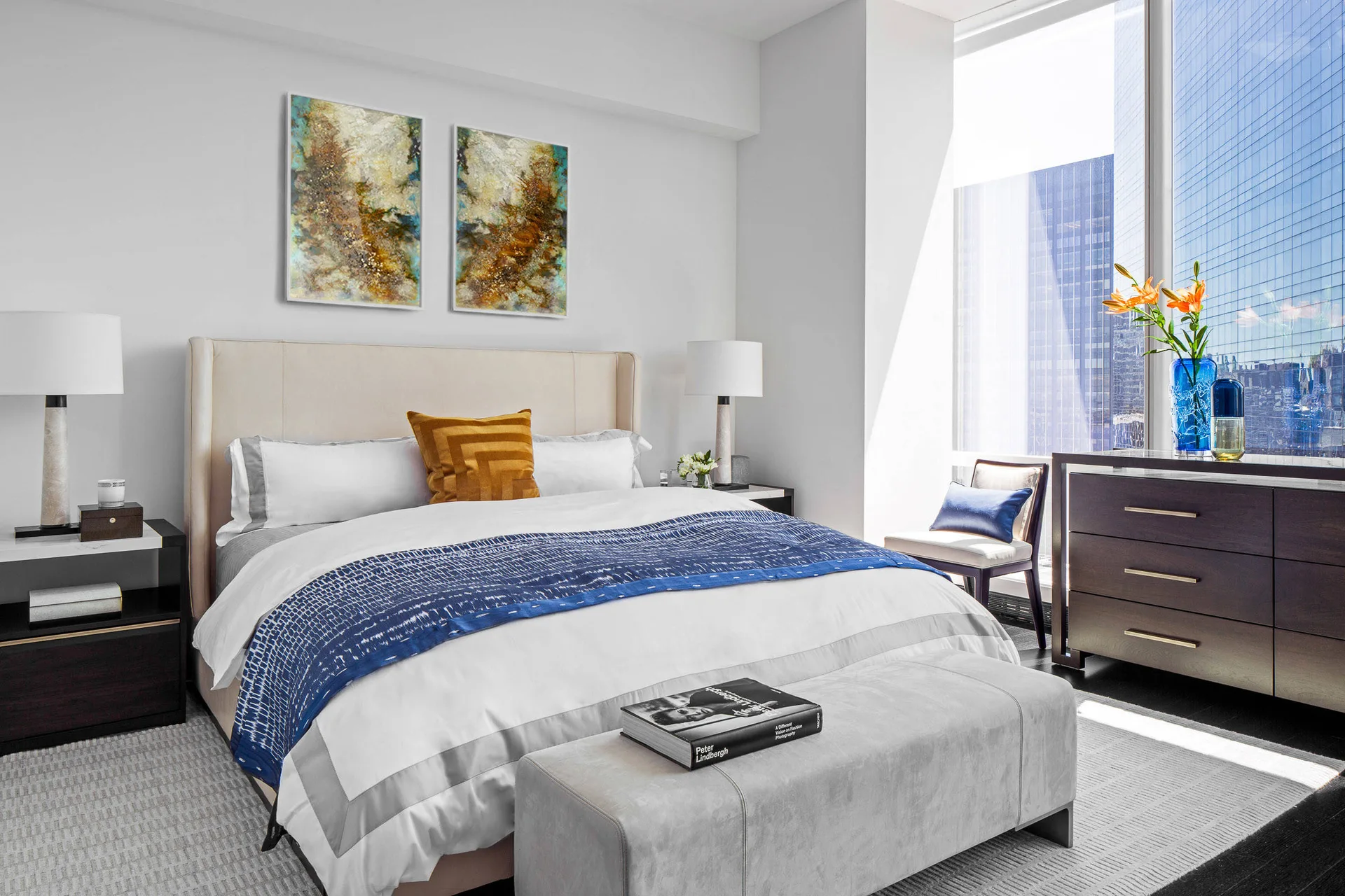 A modern bedroom features an upholstered bed with white and blue bedding, a foot bench, and a dark wood dresser next to a large window.