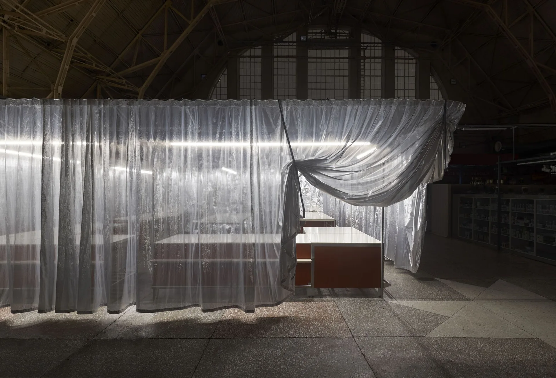 A translucent sheer white curtain encloses minimalist workstations with orange panels under bright linear lights in a large, dark industrial hall.