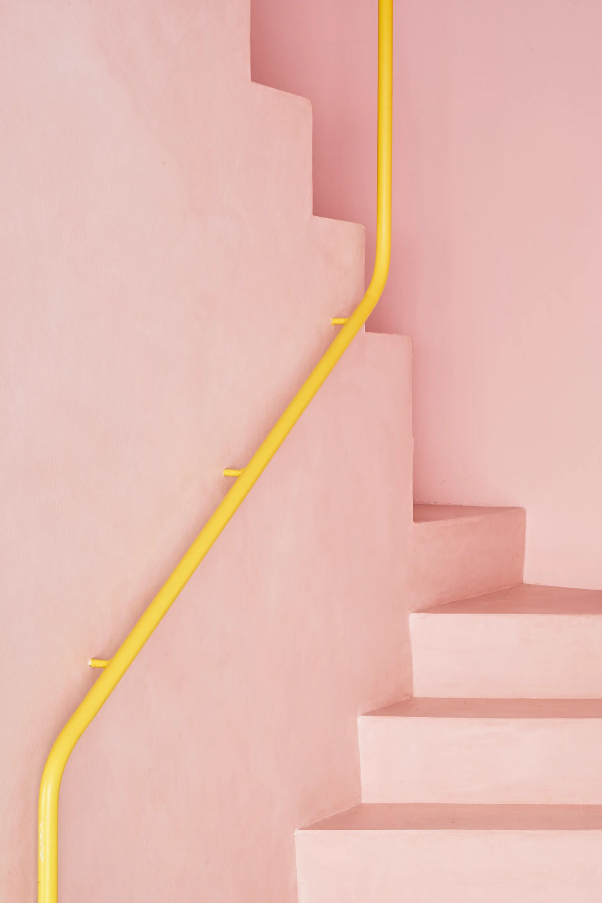 A minimalist pastel pink staircase with smooth plaster steps features a bright yellow metal tubular handrail mounted to the matching pink wall.