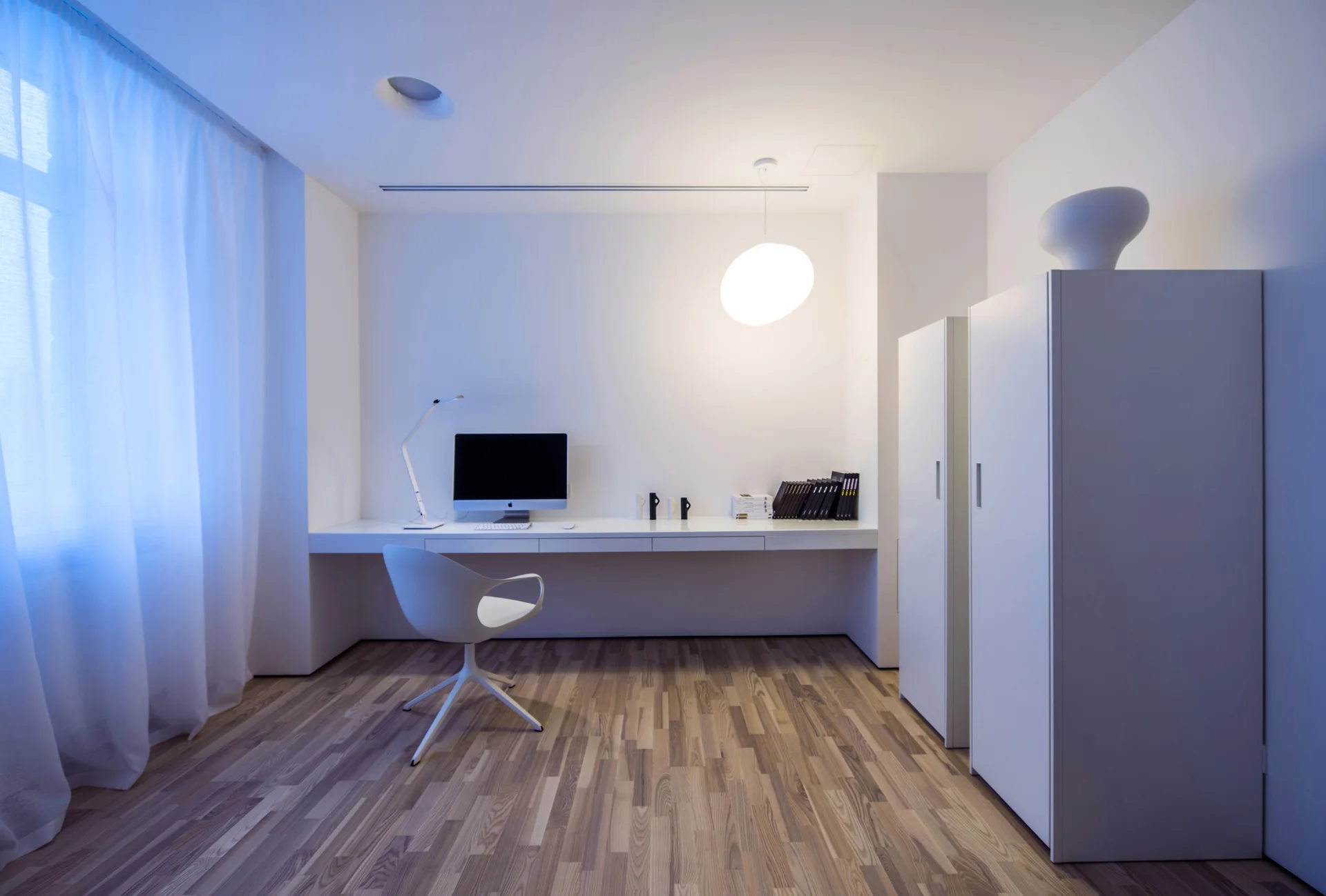 A minimalist home office features a long white built-in desk, a modern white swivel chair, tall storage cabinets, and striped light wood floors.
