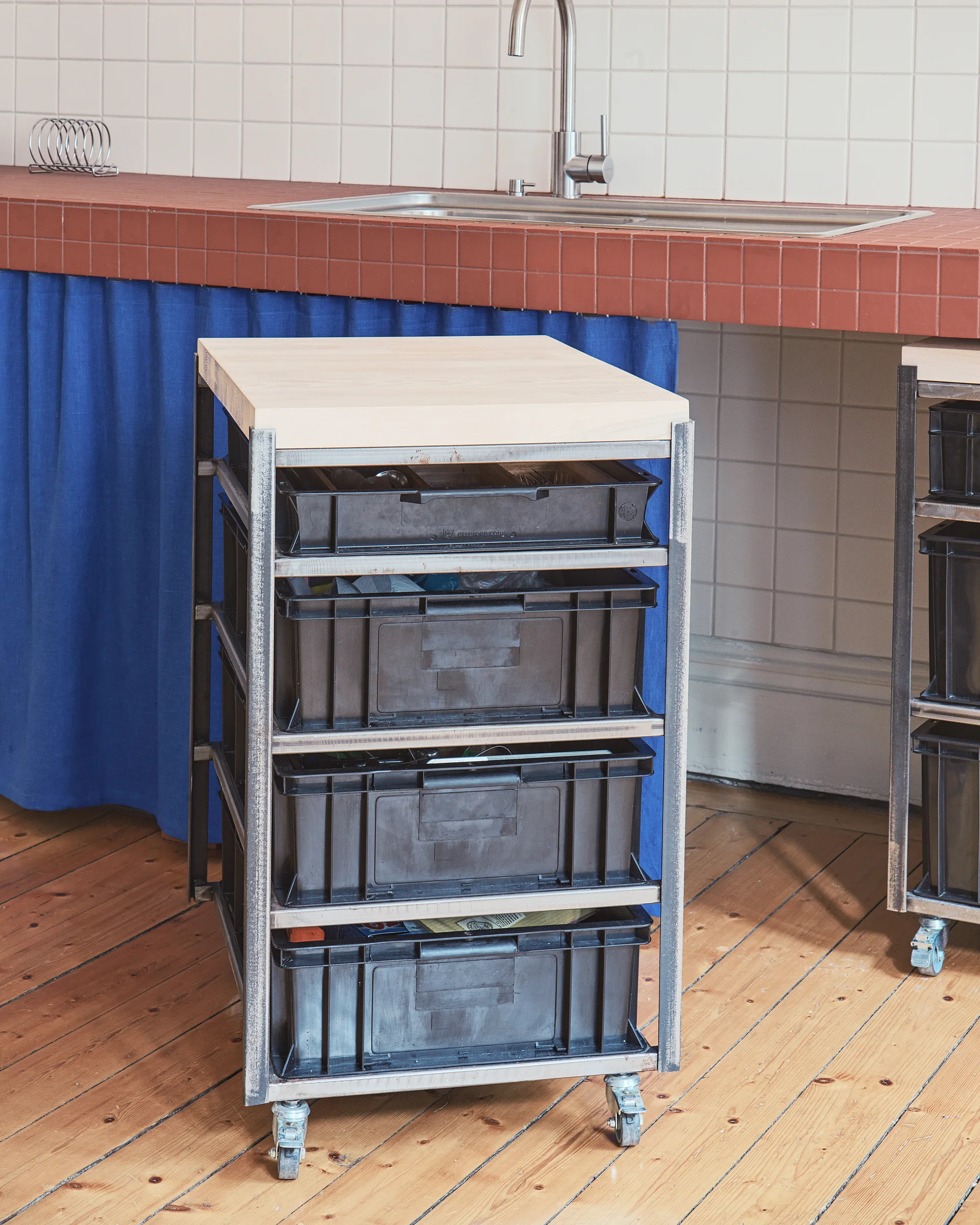 An industrial metal rolling cart with black crate drawers and a wooden top stands on wood flooring in a kitchen with tiled countertops.
