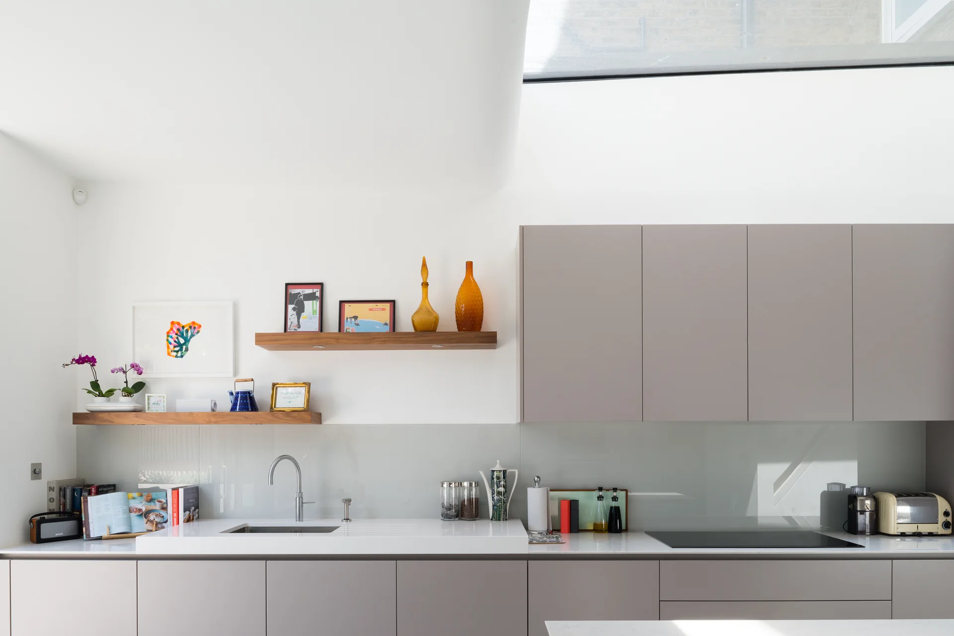 A contemporary kitchen featuring flat-front taupe cabinets, white countertops, two floating wooden shelves, and natural light from a skylight.