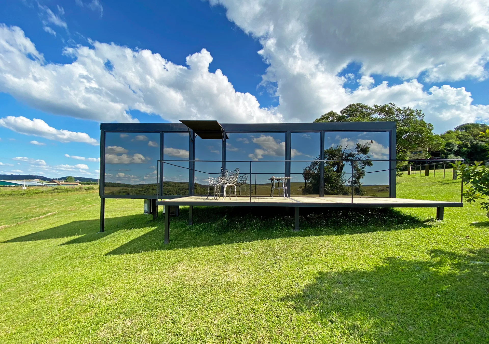 A modern black cabin with a reflective glass facade features an elevated wooden deck with metal bistro furniture set in a grassy field.