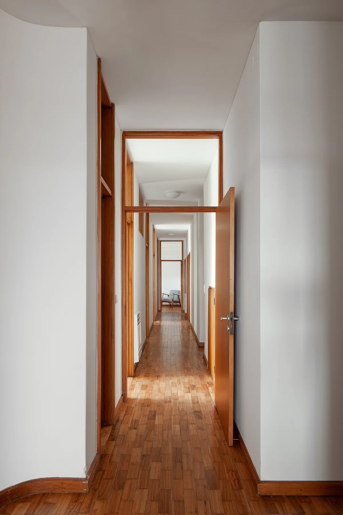 A long hallway features matte white walls, matching wooden door frames, and parquet block flooring stretching into the distance.