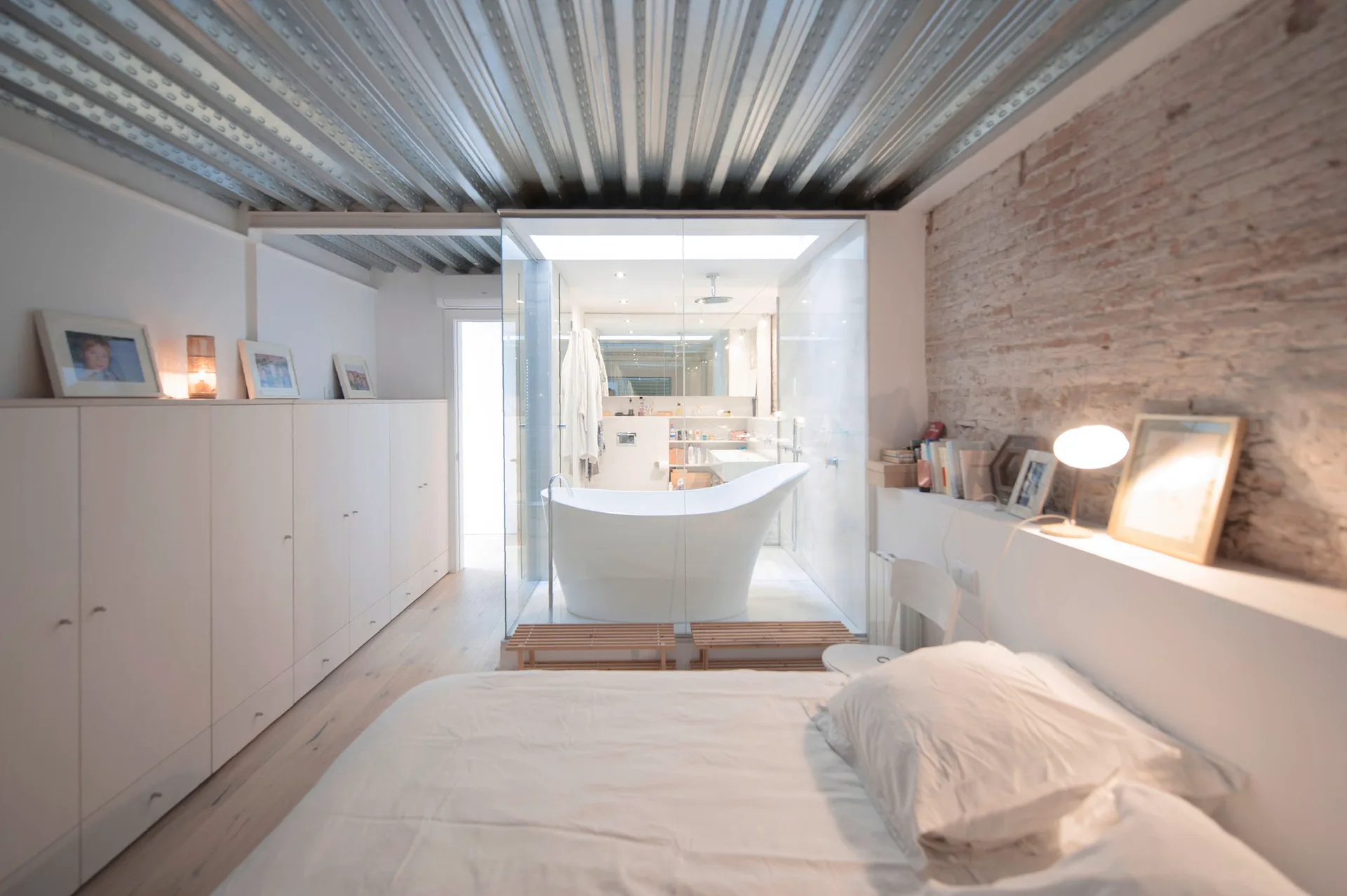 An industrial bedroom featuring a corrugated metal ceiling, an exposed brick wall, white built-in cabinets, and a glass-enclosed central bathroom.