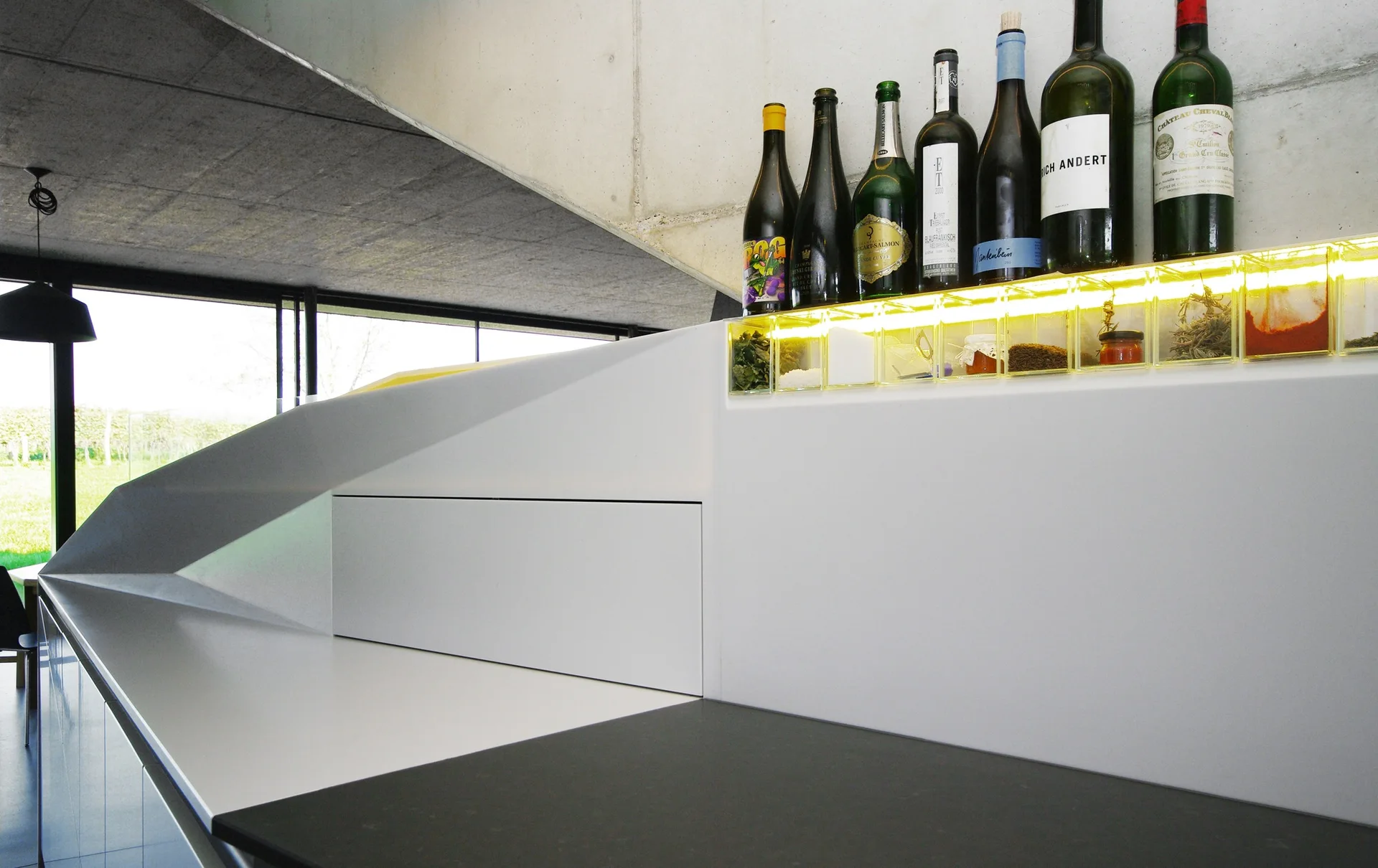 A modern kitchen counter features a row of wine bottles above a glowing, backlit spice storage compartment with an exposed concrete ceiling overhead.