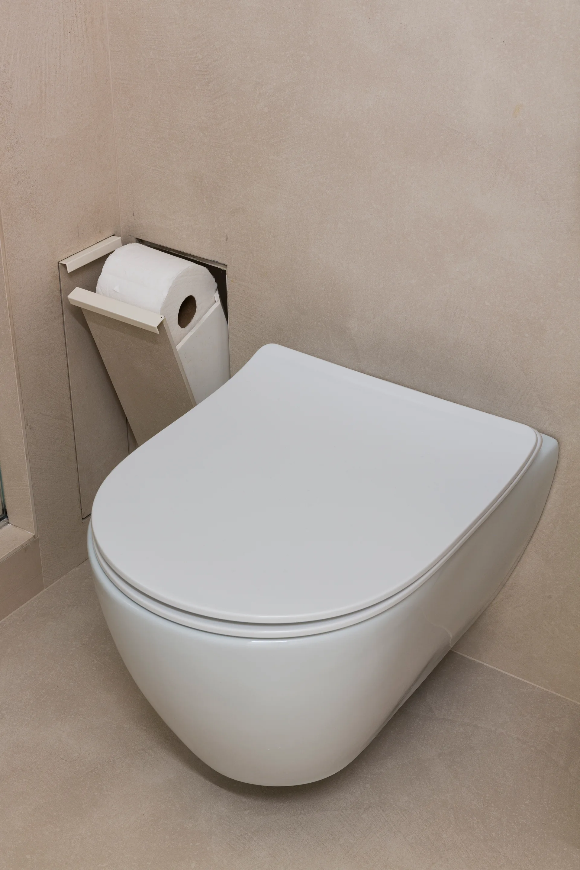A sleek white wall-mounted toilet is positioned next to a matching recessed toilet paper holder set into beige tiled walls.