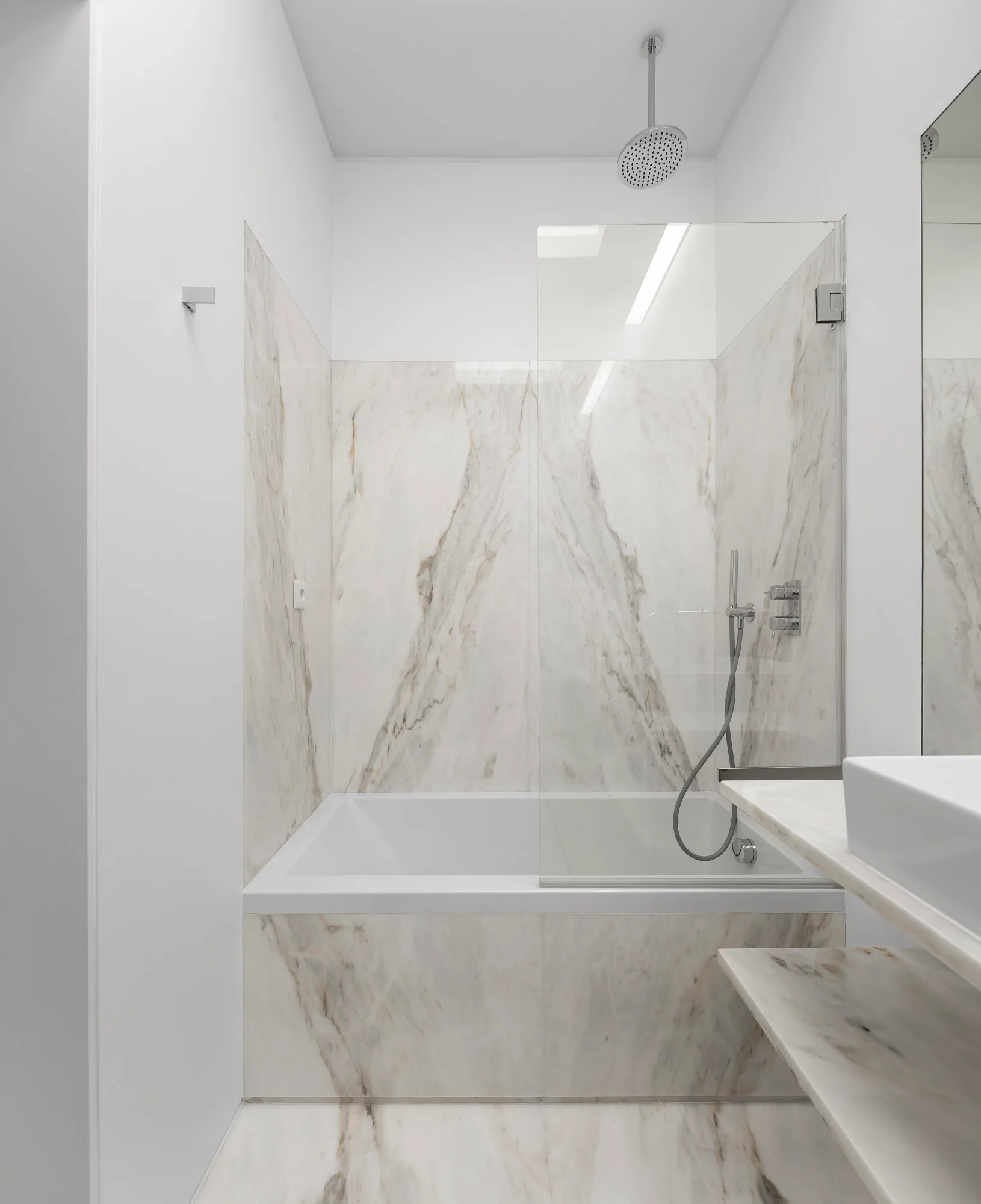A modern white bathroom features a built-in bathtub enclosed in veined marble slabs with a frameless clear glass shower panel.