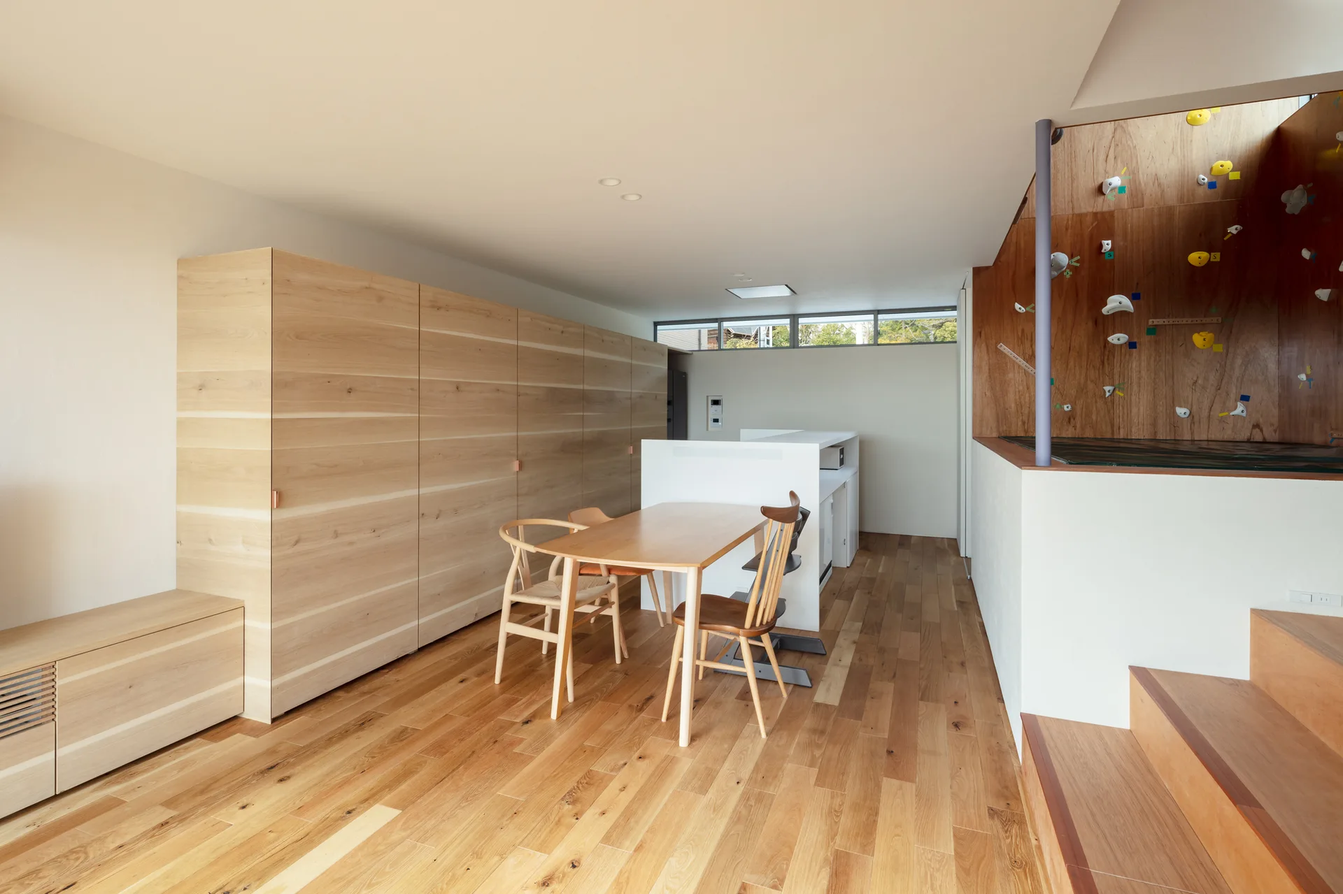 An open-plan dining room features light wood floors, a matching dining set, a wall of built-in wooden cabinets, and a climbing wall in the background.