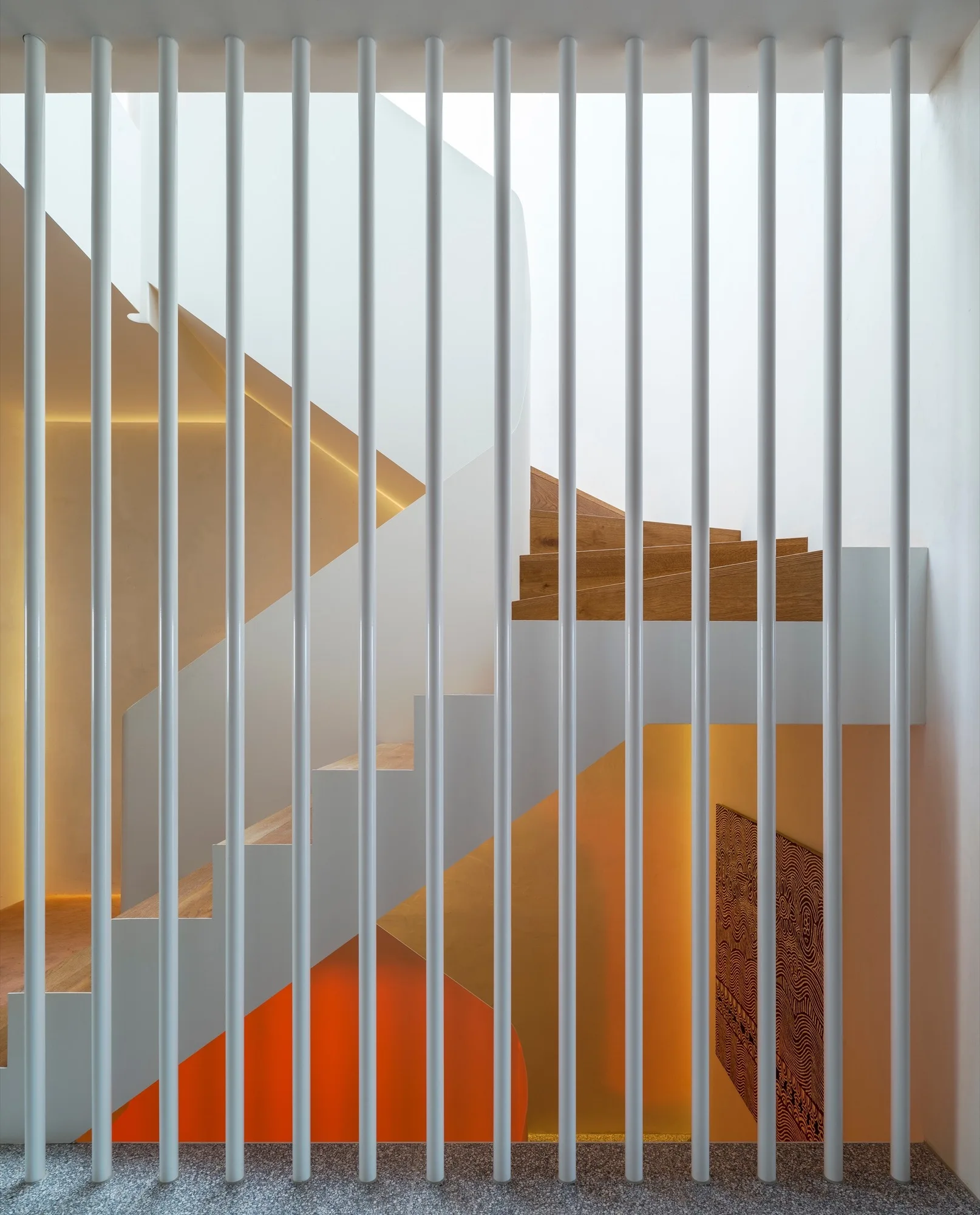 A modern staircase features light wood treads behind a screen of floor-to-ceiling white vertical metal rods with warm glowing accent lighting on the walls.
