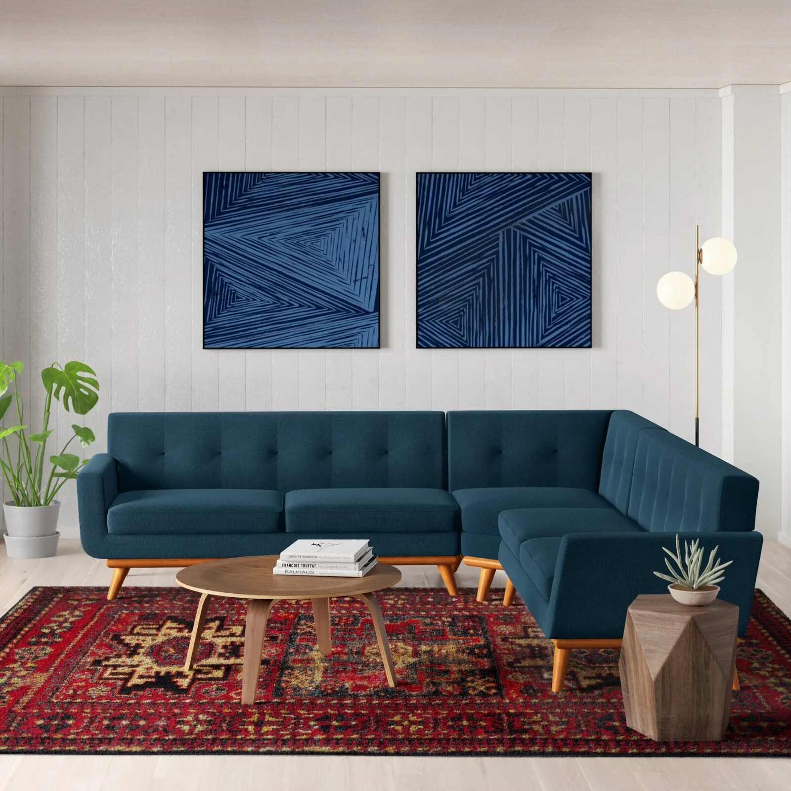 A blue sectional sofa sits on a red patterned rug in front of a white vertical paneled wall decorated with two blue abstract art prints.