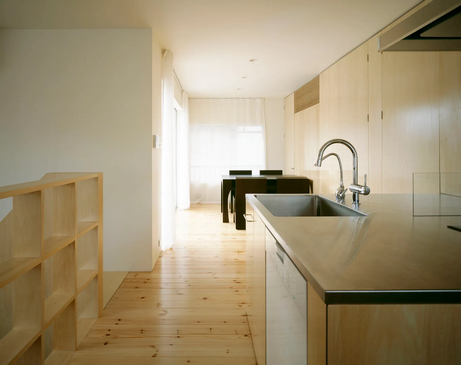 A minimalist kitchen features a stainless steel countertop with a sink, light wood cabinetry, and matching wide-plank wood floors.