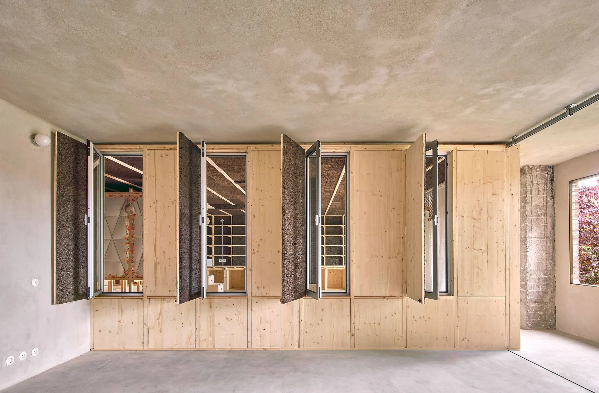 A minimalist room featuring light pine wood wall paneling with open integrated shutters alongside a raw concrete floor and ceiling.