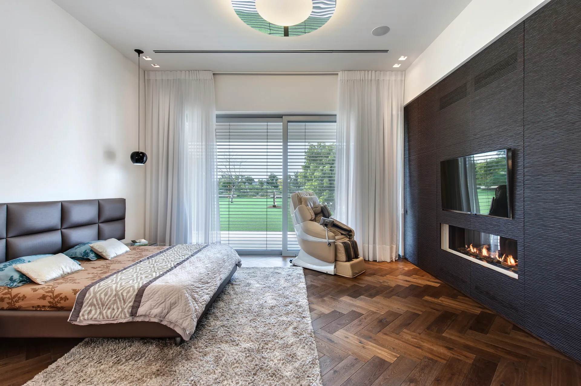 A modern bedroom featuring a dark textured media wall with an integrated fireplace, a low-profile upholstered bed, and herringbone wood flooring.