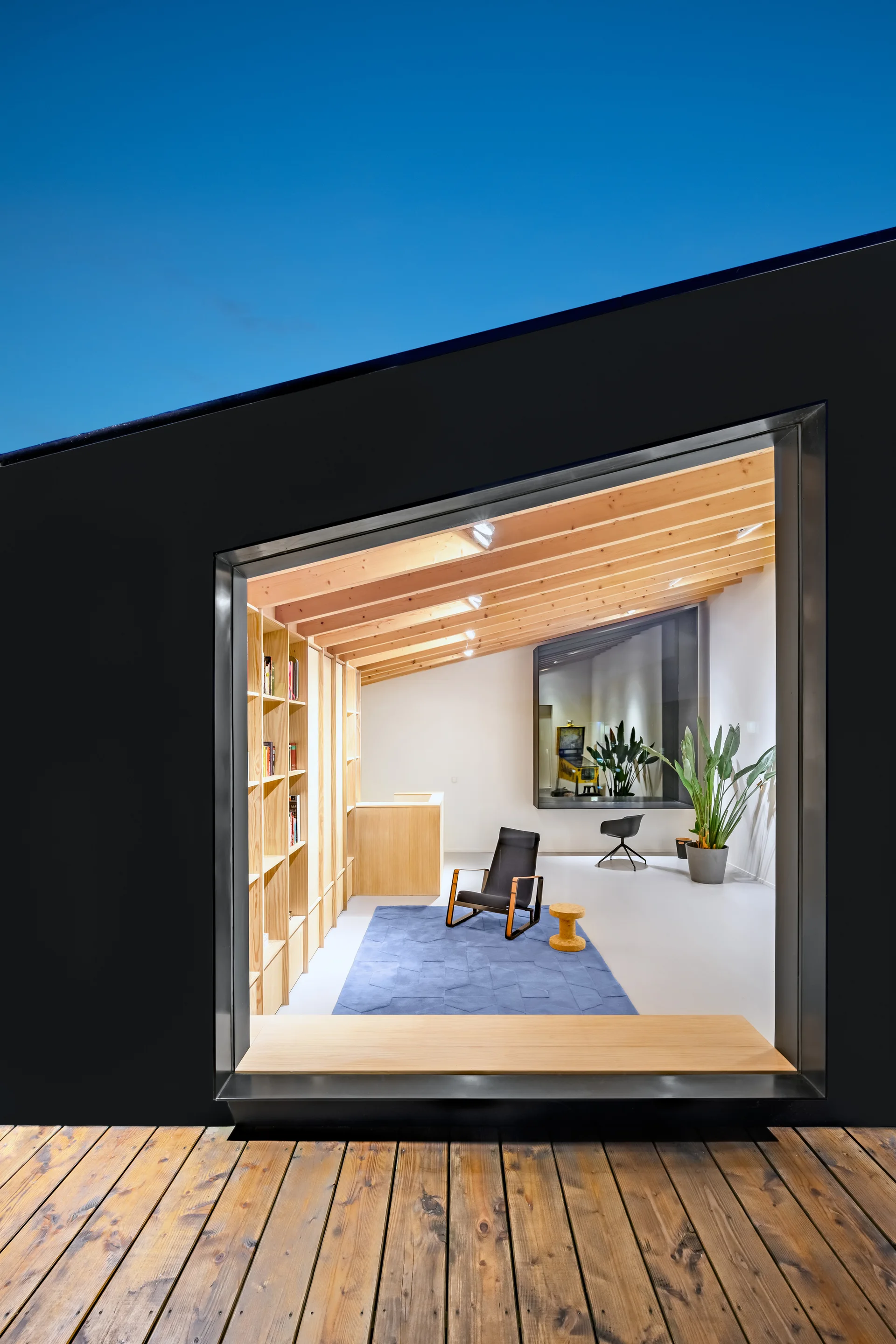 An interior room with light wood ceiling beams and built-in bookshelves is viewed through a large window from an outdoor wooden deck.