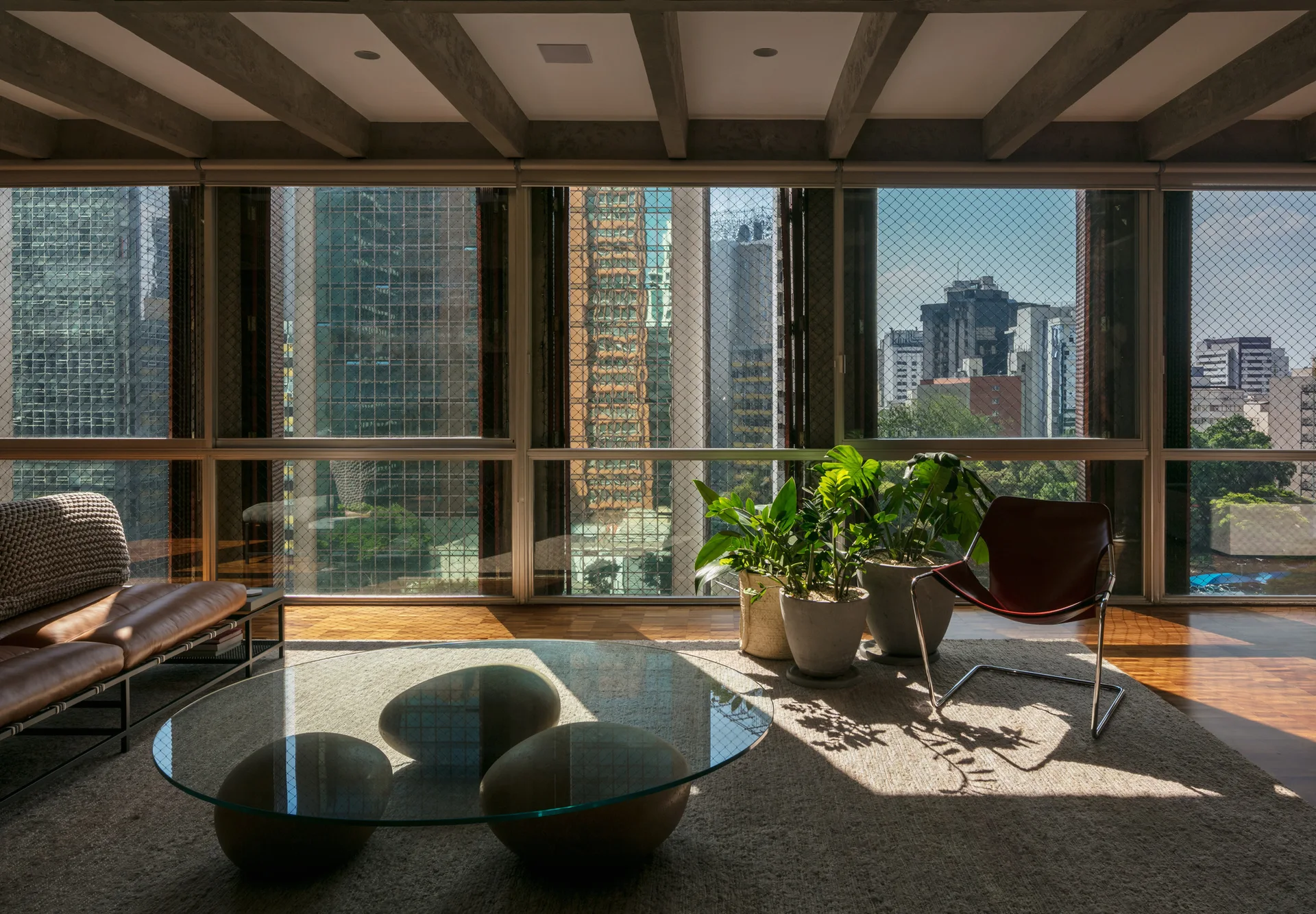 A modern living room features exposed concrete ceiling beams, floor-to-ceiling windows showing a city view, a glass coffee table, and leather seating.