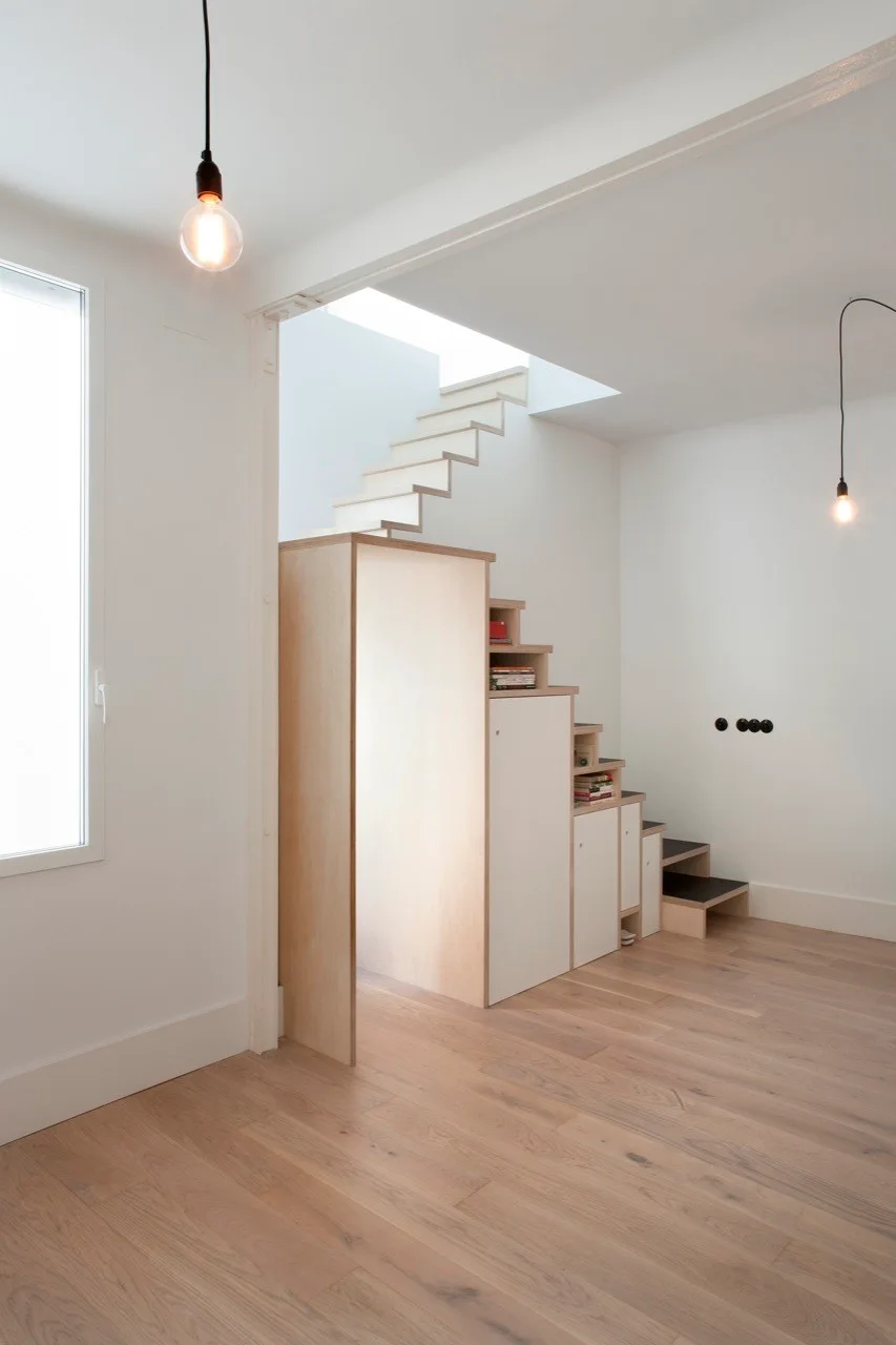 A minimalist plywood staircase features integrated white cabinets and open shelving on a light hardwood floor.