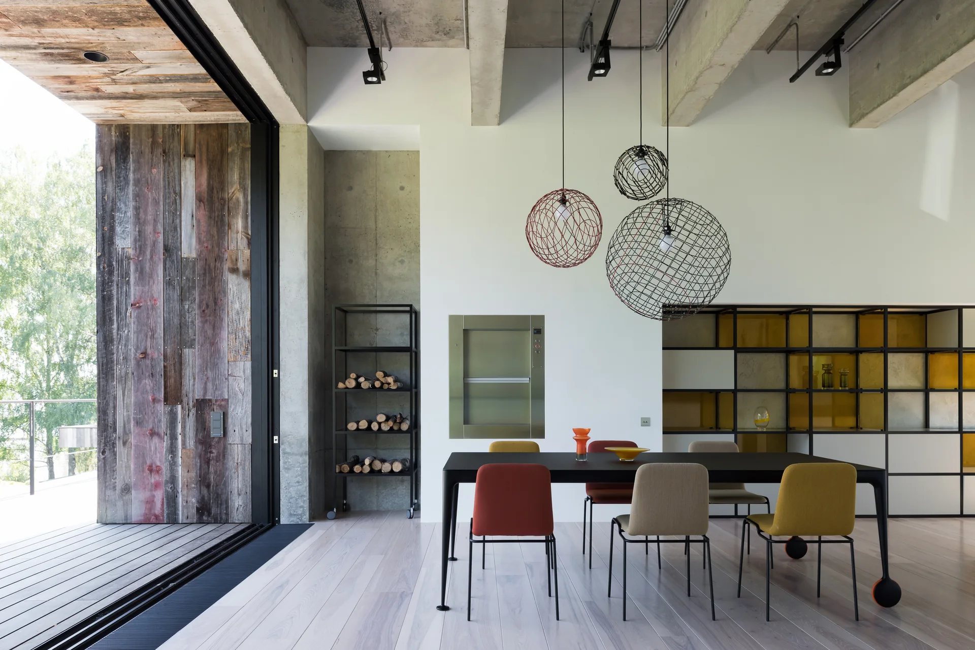 A modern dining room features a black table with colorful chairs beneath exposed concrete ceiling beams and three spherical wire pendant lights.