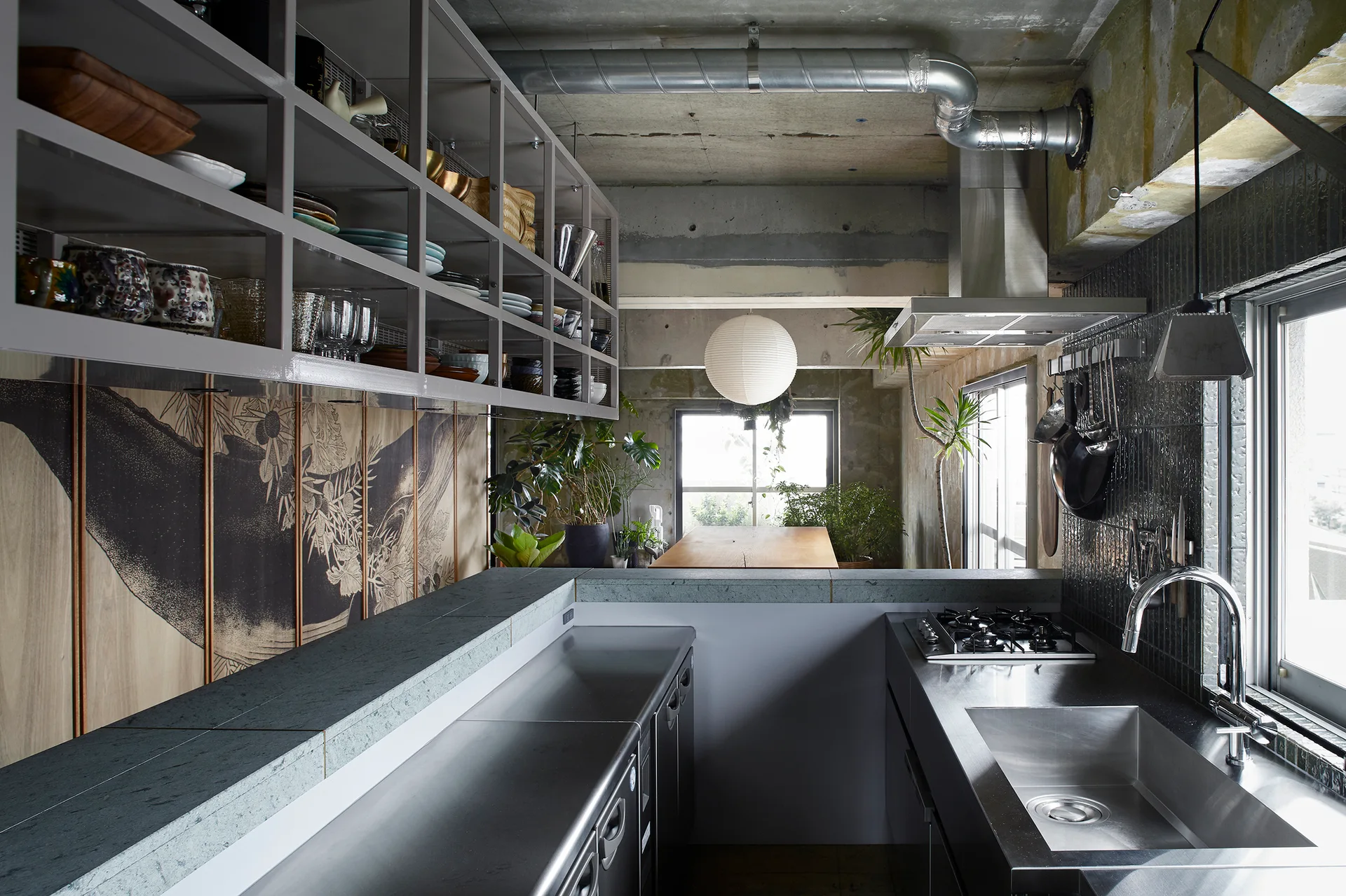 An industrial galley kitchen featuring stainless steel countertops, a raw concrete ceiling with exposed ductwork, and suspended grey grid shelving.