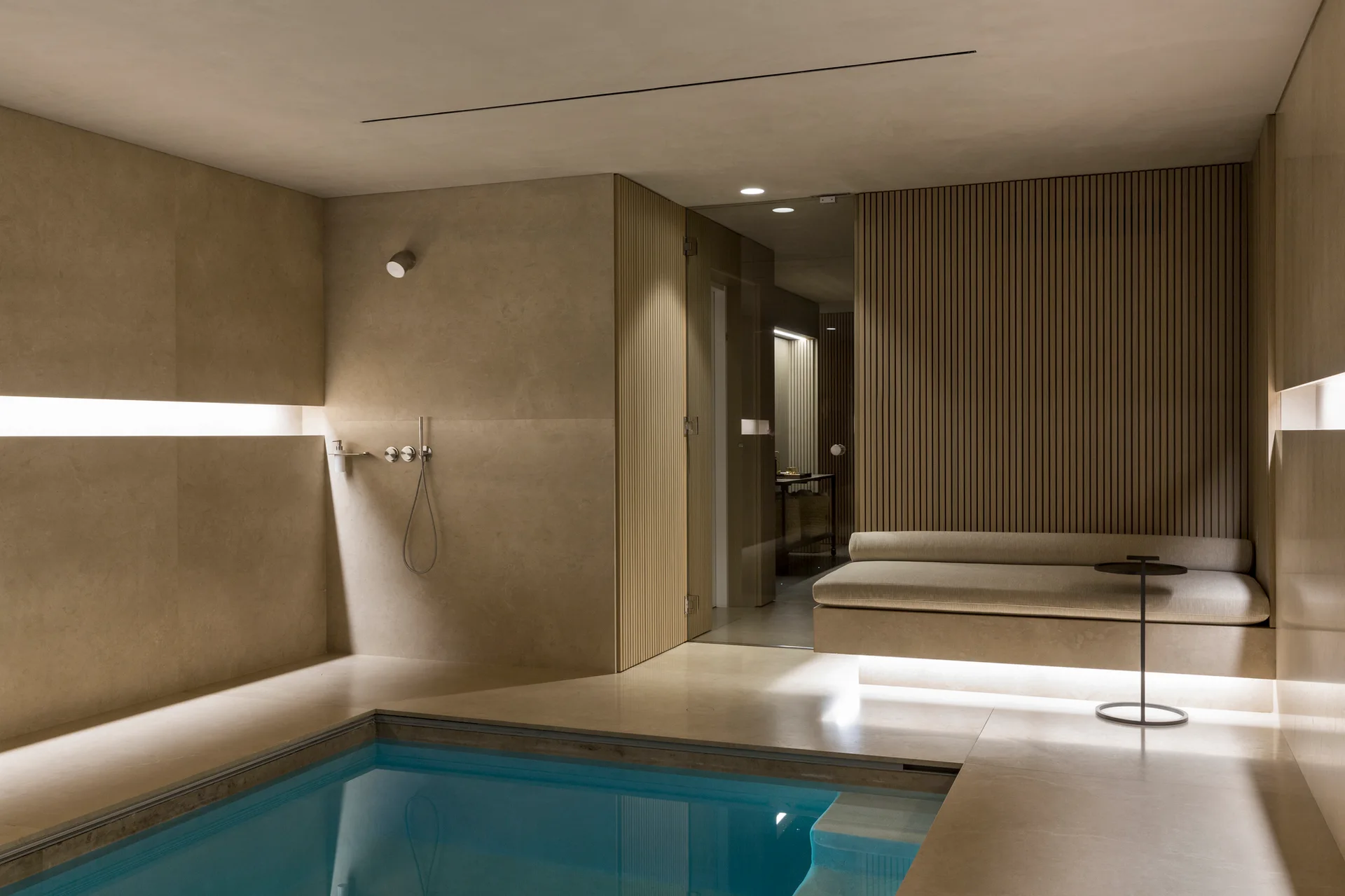 A minimalist indoor spa featuring a blue swimming pool, beige stone tile walls, a built-in cushioned bench, and vertical wood slat paneling.