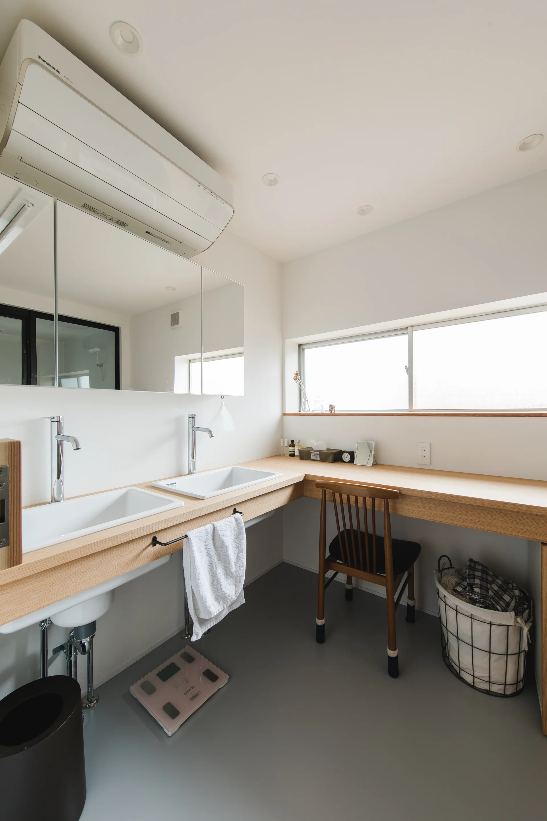 A minimalist bathroom features an L-shaped light wood vanity with two sinks, a wooden spindle chair, grey flooring, and a wide wall mirror.