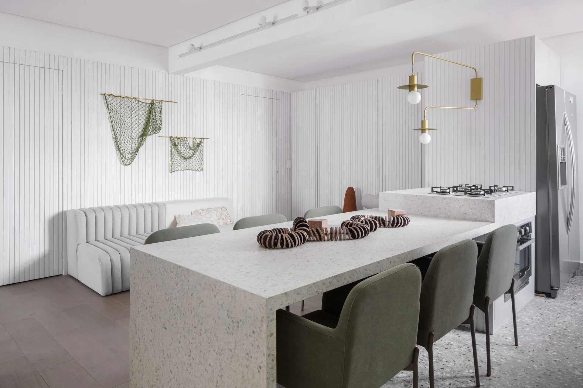 A modern kitchen featuring a speckled white terrazzo island with olive green chairs, a brass sconce, and a white slatted accent wall.