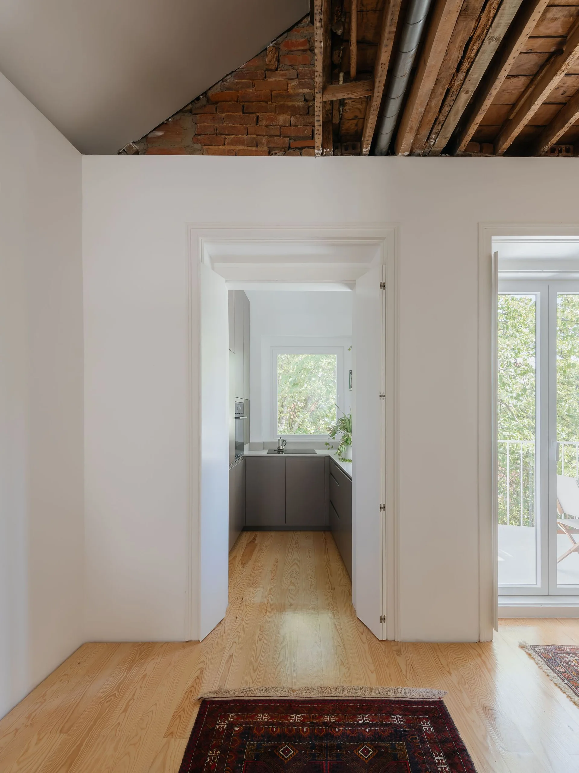 A view through a white doorway into a kitchen with gray cabinets, showing an exposed brick and timber ceiling overhead and light wood floors.