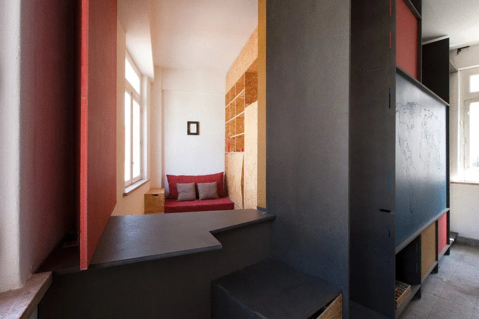 A modern studio room featuring a large dark grey wooden partition, a raised platform, and a small alcove containing a red sofa and raw OSB shelving.