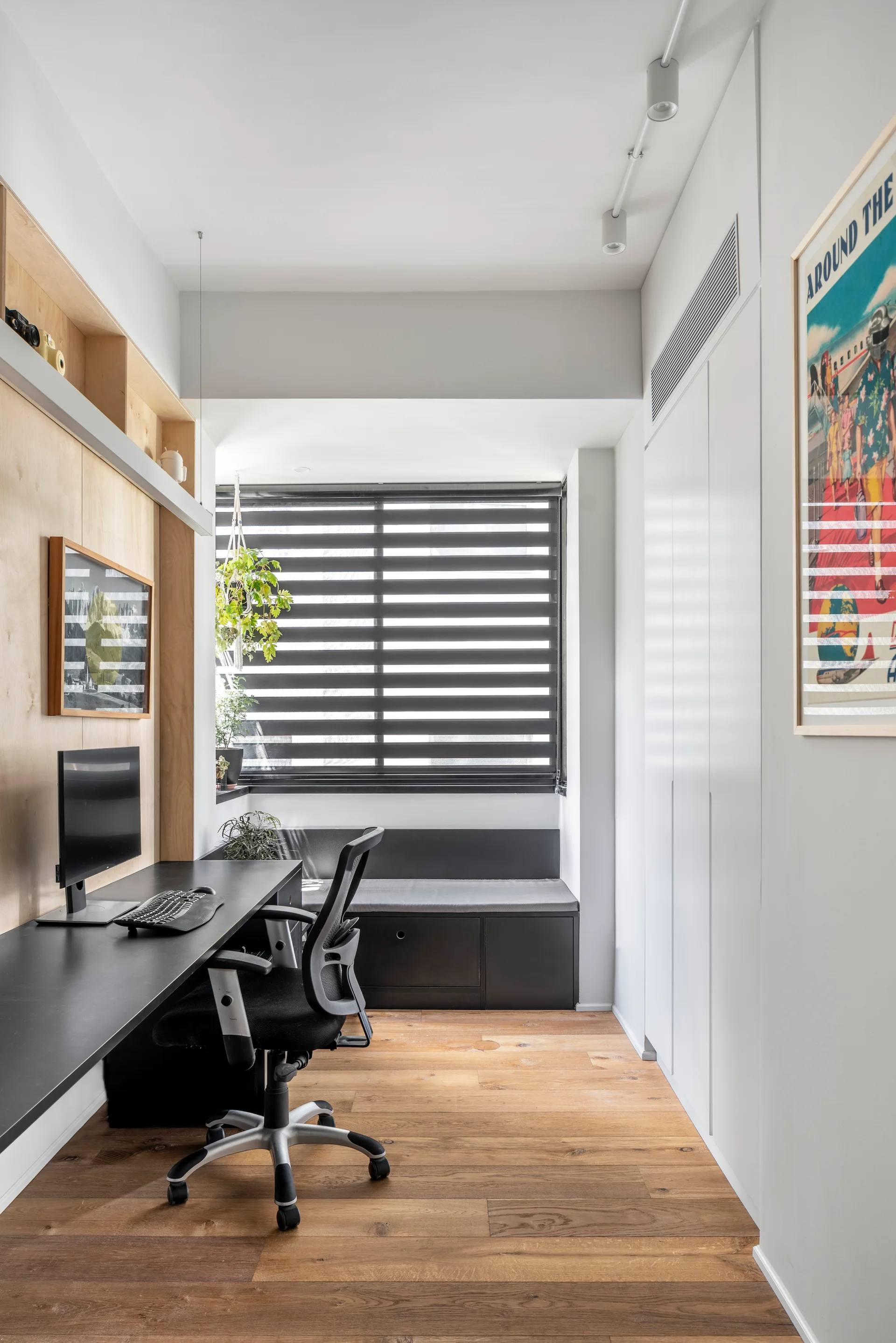 A narrow home office with a black desk, wood plank flooring, a built-in window seat with striped blinds, and floor-to-ceiling white cabinets.