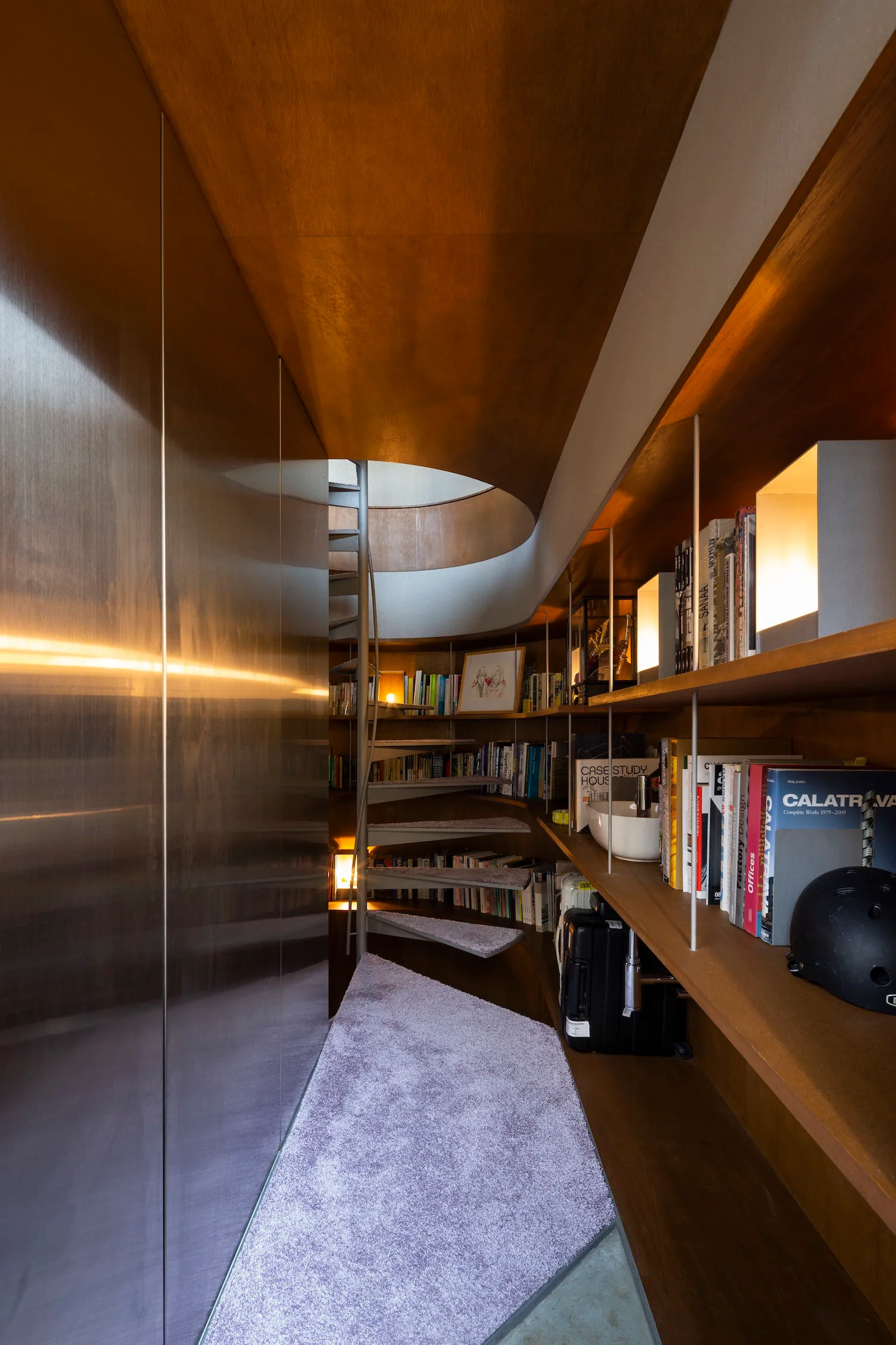 A compact spiral staircase sits between integrated curved wooden bookshelves and a large brushed metal wall panel under a wood ceiling.