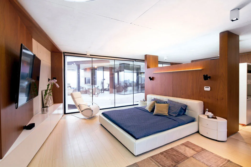 A modern bedroom features a low-profile white bed with blue bedding, a wood-paneled partition wall, and large floor-to-ceiling glass windows.
