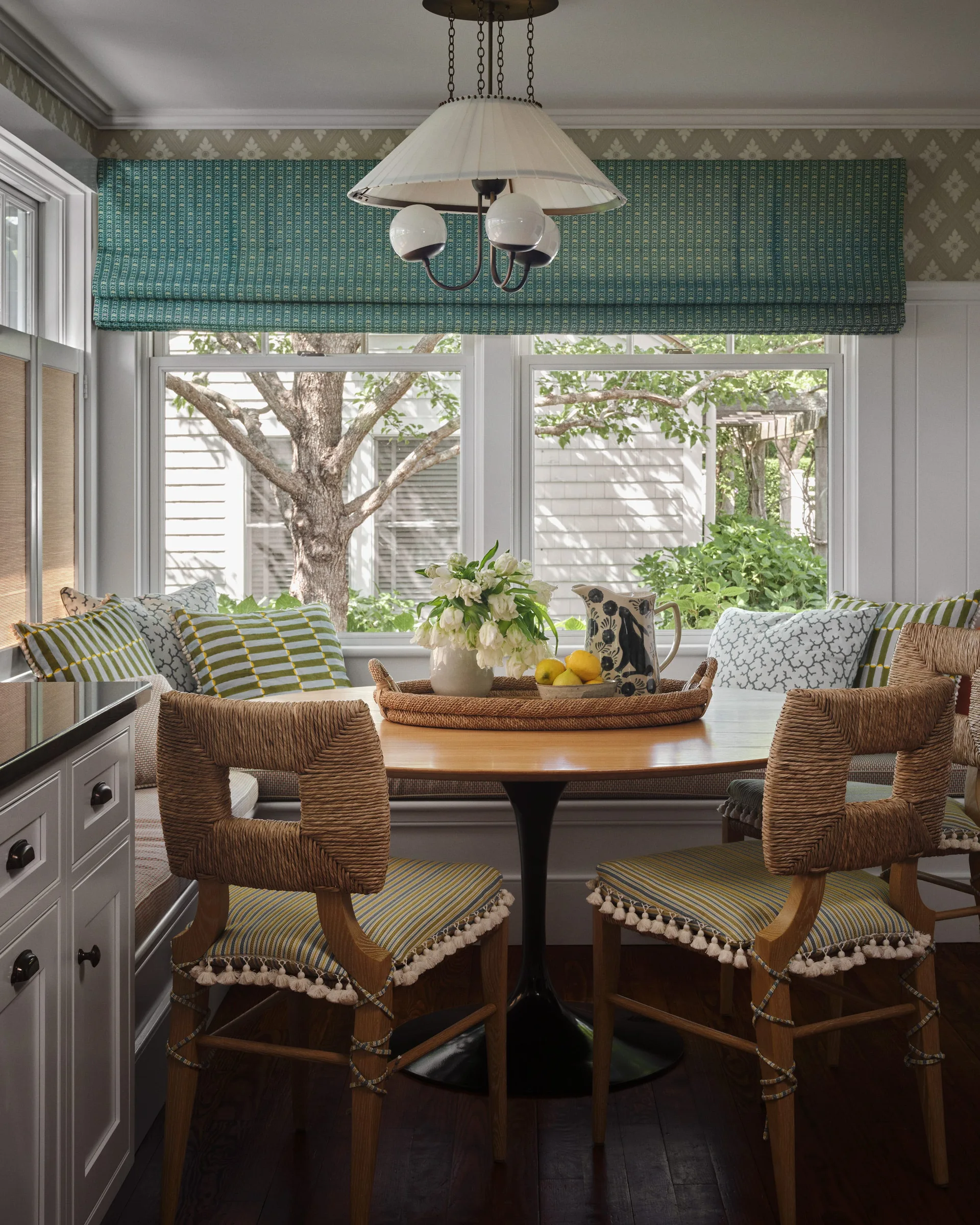 A bright breakfast nook features a circular wooden table surrounded by woven wood chairs with tassel trim and a built-in window banquette.
