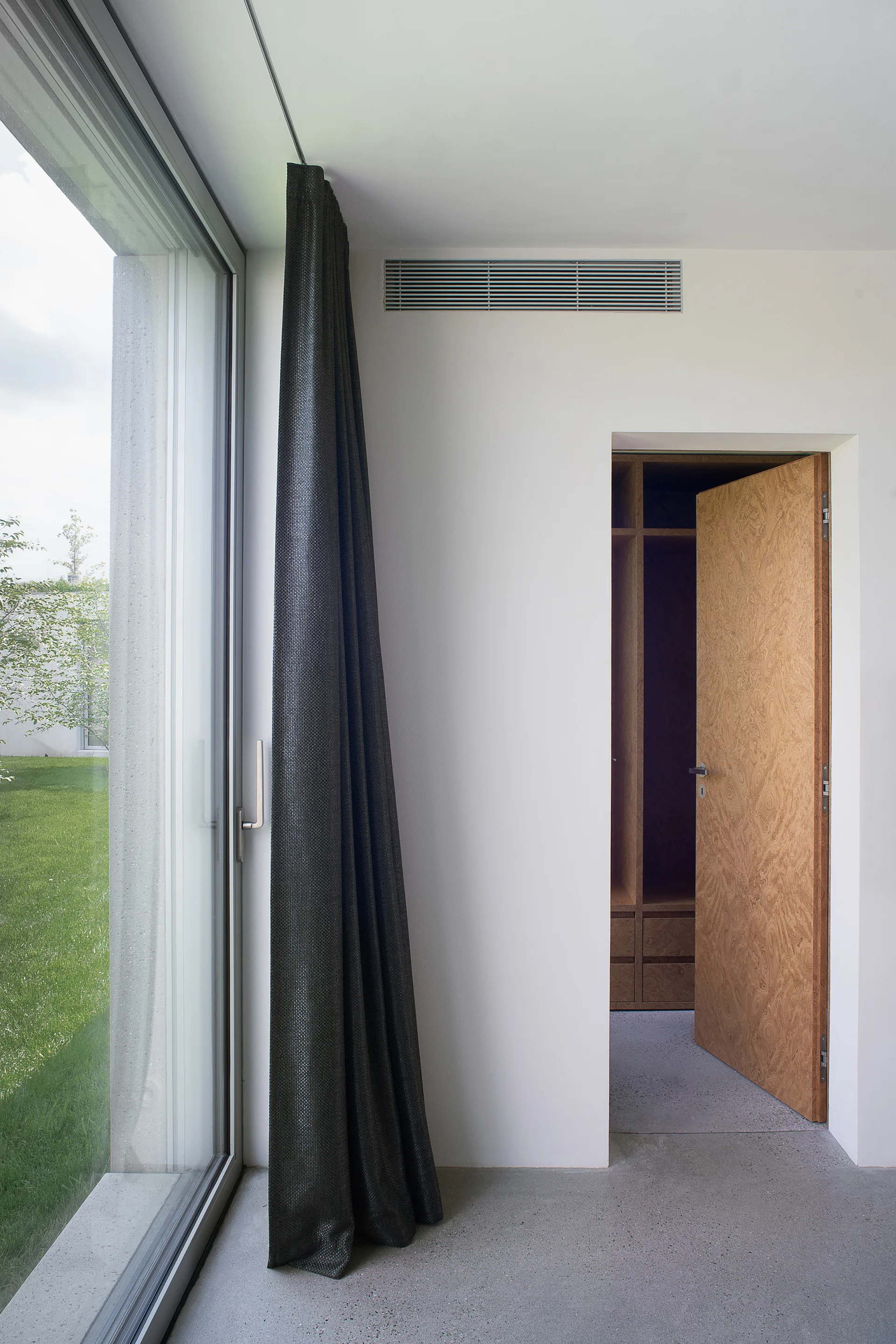 An open textured wooden door reveals a walk-in closet next to a white wall with a dark curtain hanging beside a large glass window.