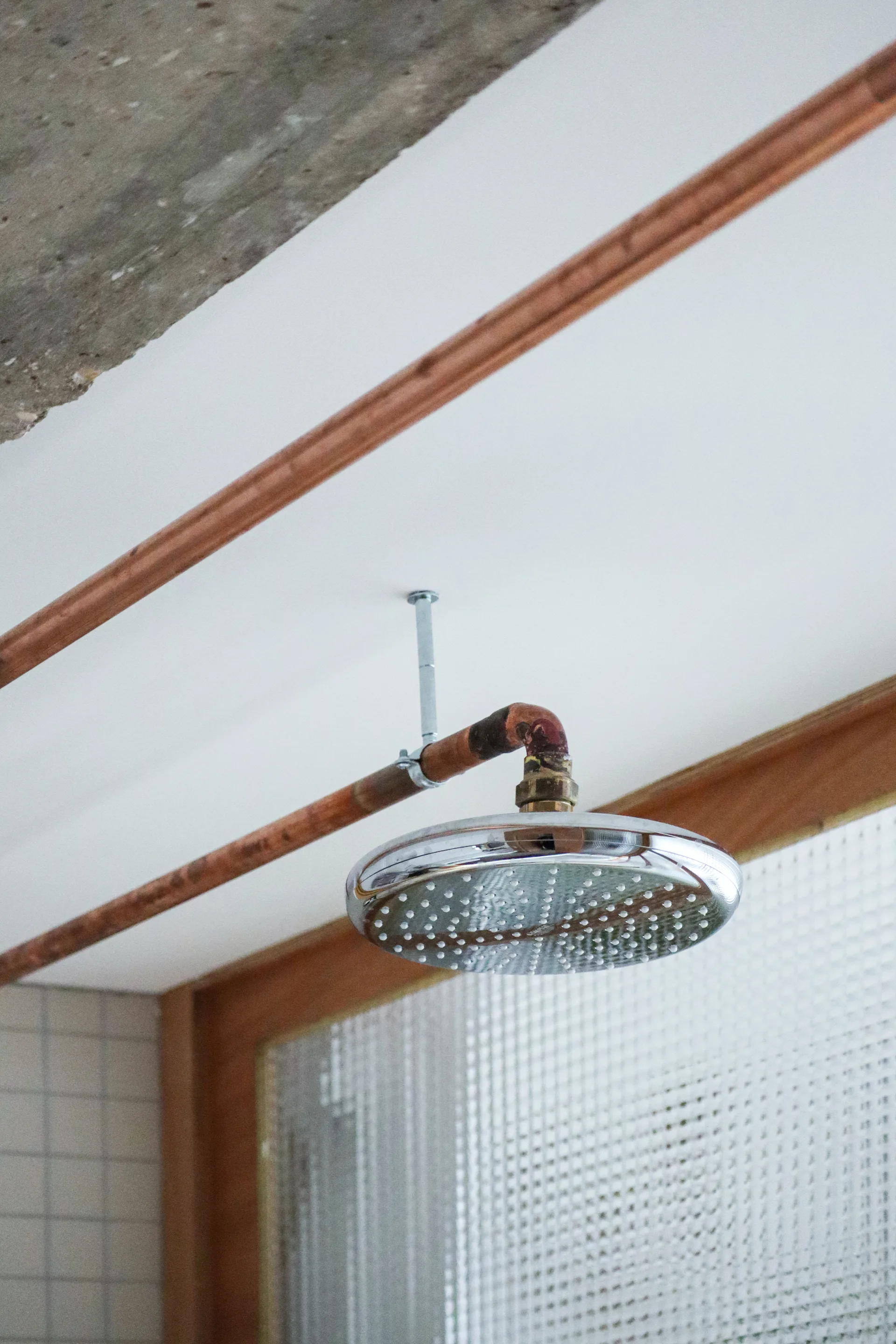 A circular chrome shower head is mounted to exposed copper pipes against a white ceiling and textured glass partition.