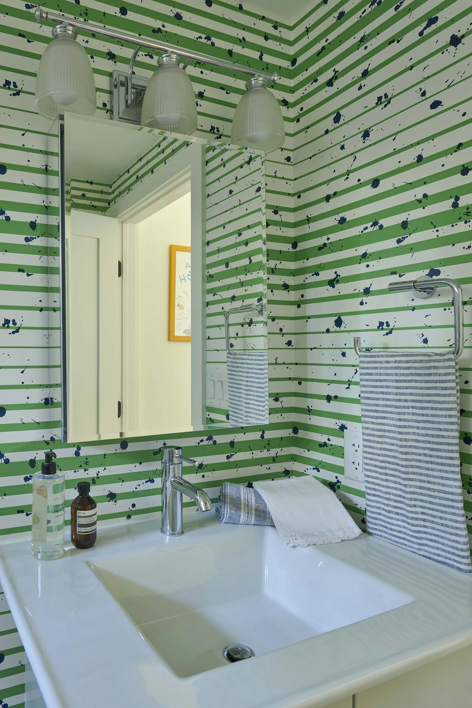 A bathroom vanity features green and white striped wallpaper with blue paint splatters, a white undermount sink, and chrome fixtures.