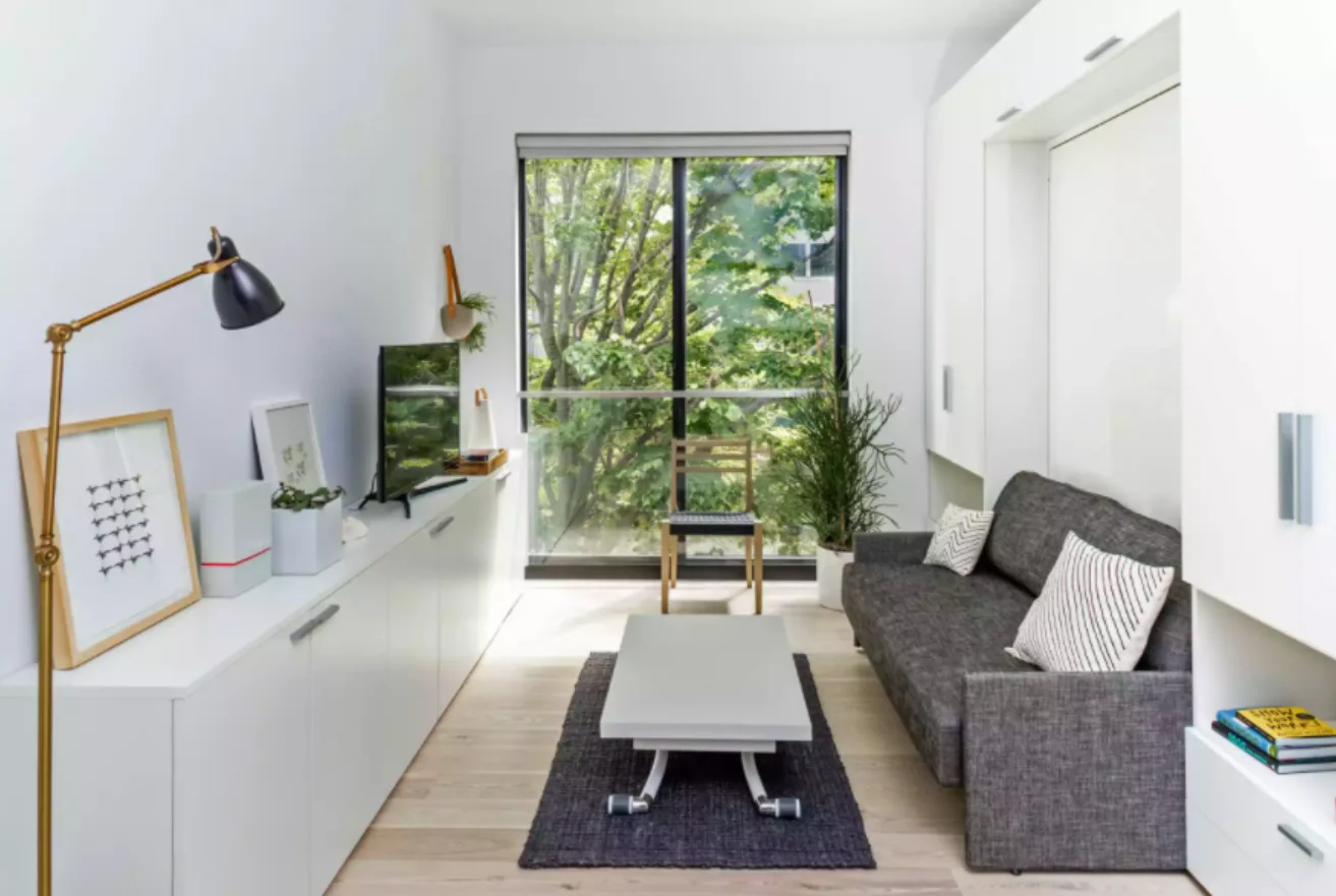 A narrow living room features a grey sofa, a low coffee table, and long white built-in storage cabinets opposite a large window.